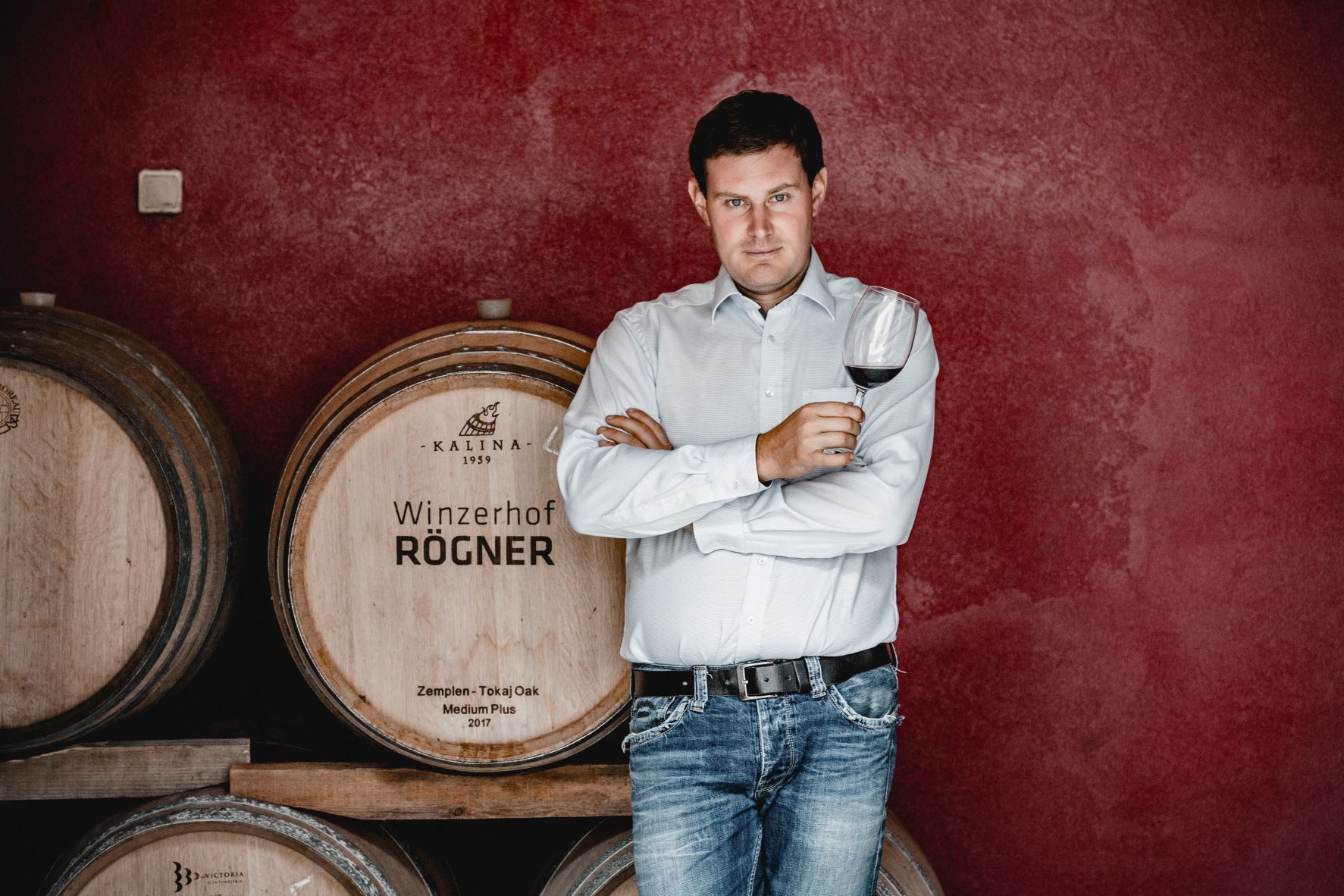 Ein Mann steht vor Weinfässern und hält ein Glas Rotwein in der Hand.