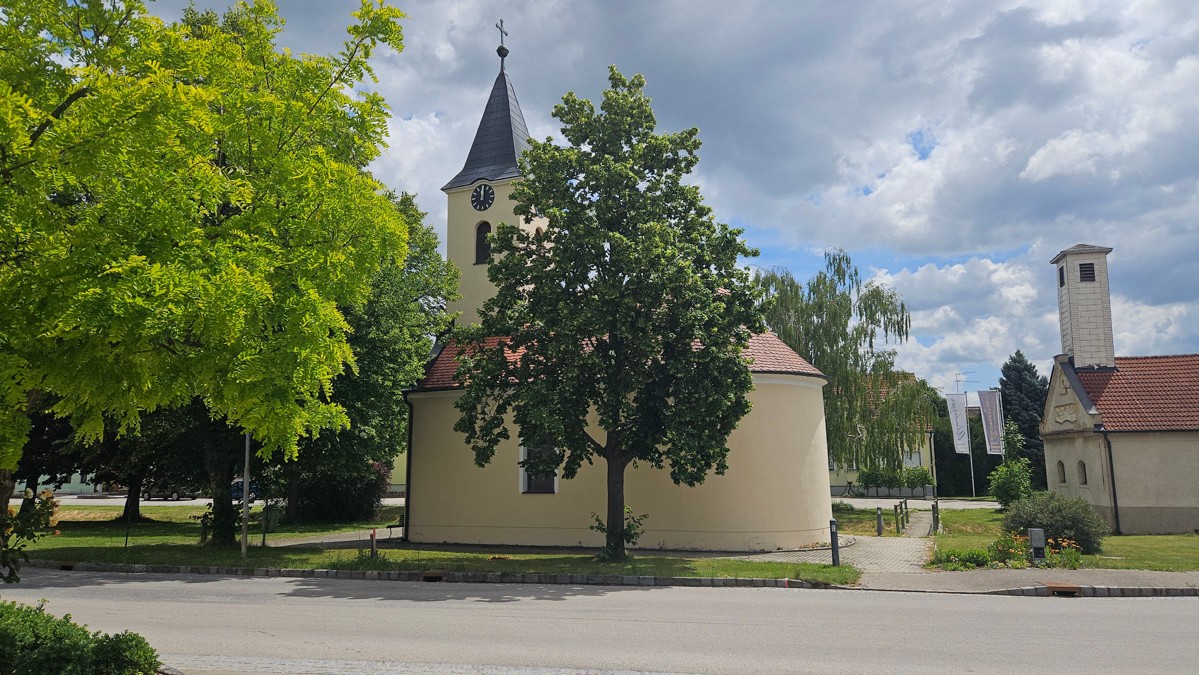 Eine kleine Kapelle mit einem Turm und einer Uhr, umgeben von Bäumen und einem blauen Himmel mit Wolken.