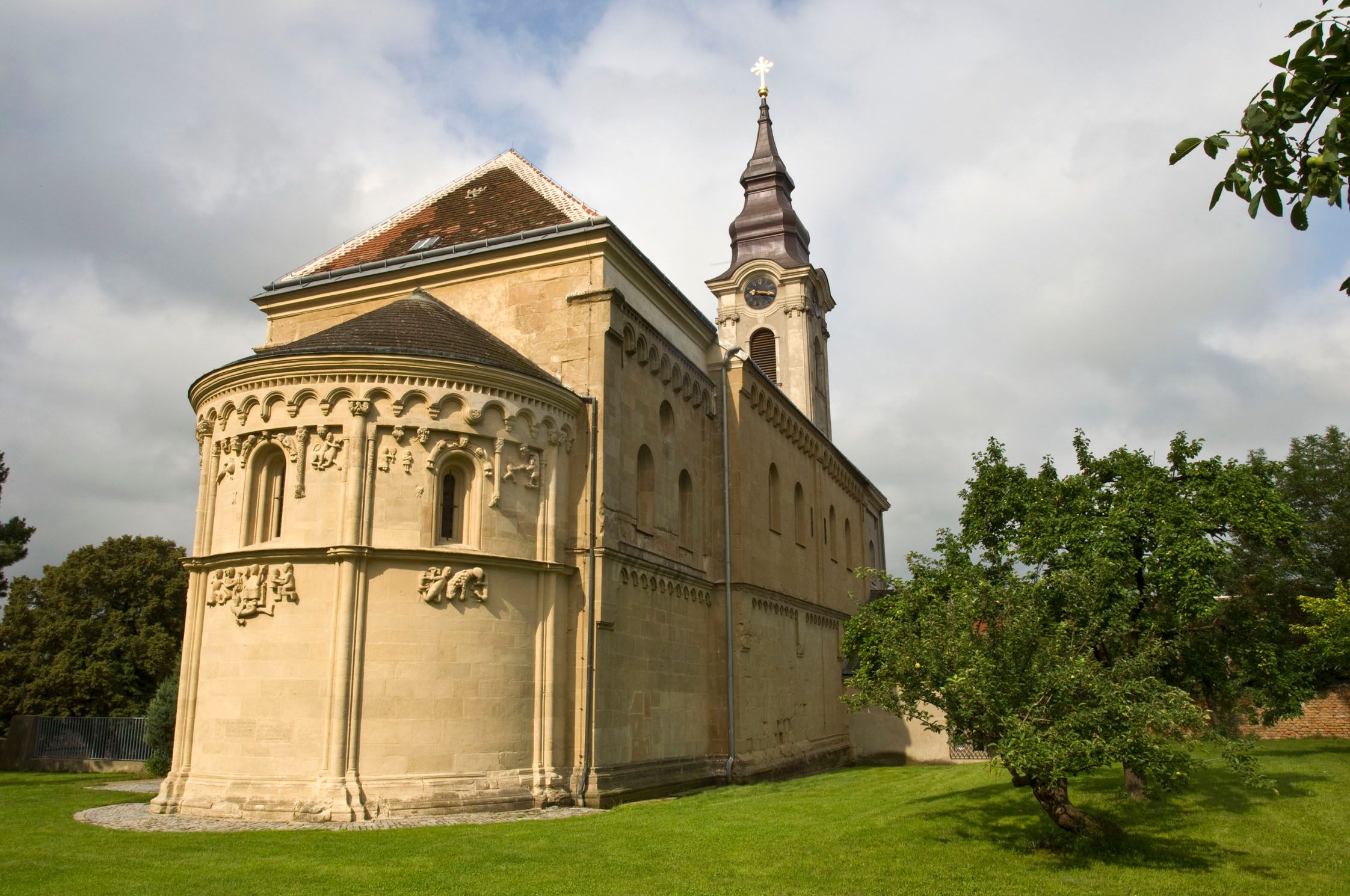 Romanische Kirche mit Verzierungen und Turm, umgeben von grüner Wiese und Bäumen.