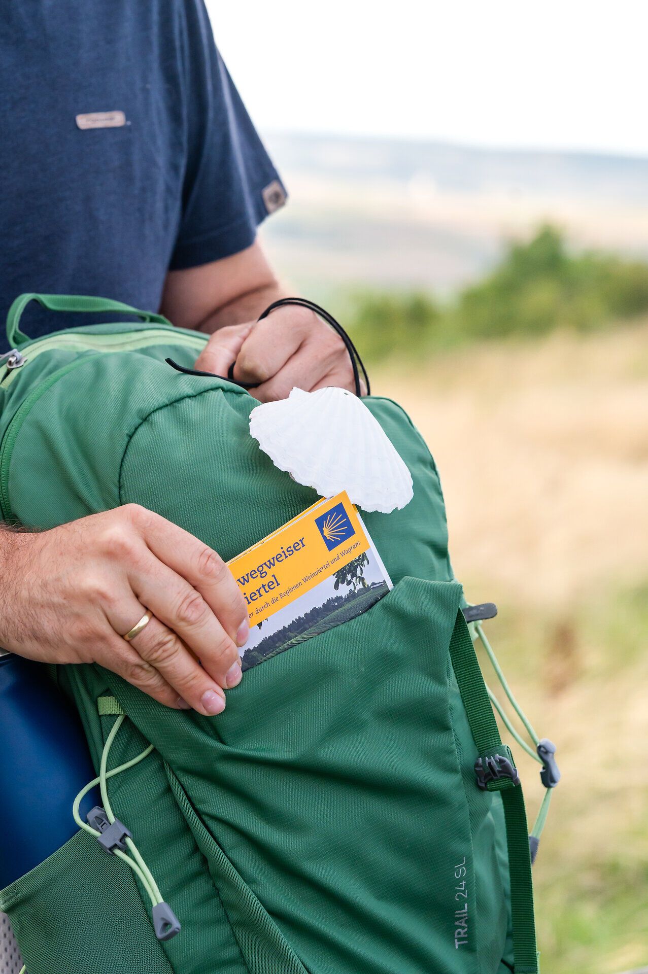 Eine Person greift mit der Hand in ein Fach eines Wanderrucksackes und zieht ein Reisehandbuch zum Jakobsweg Weinviertel heraus. Auf dem Rucksack ist eine Jakobswegmuschel befestigt.