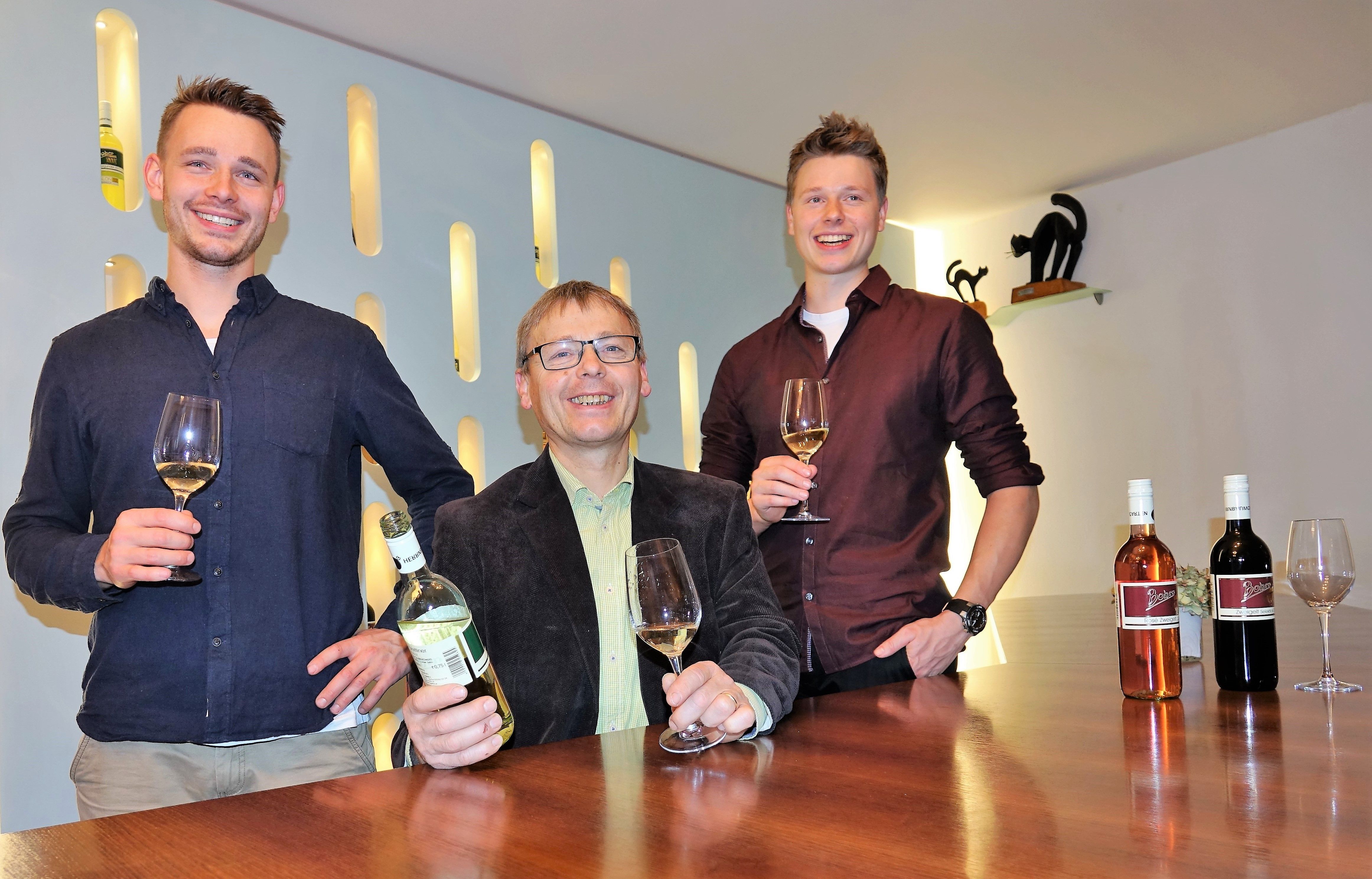 Three men are standing at a table with wine glasses and bottles of wine.
