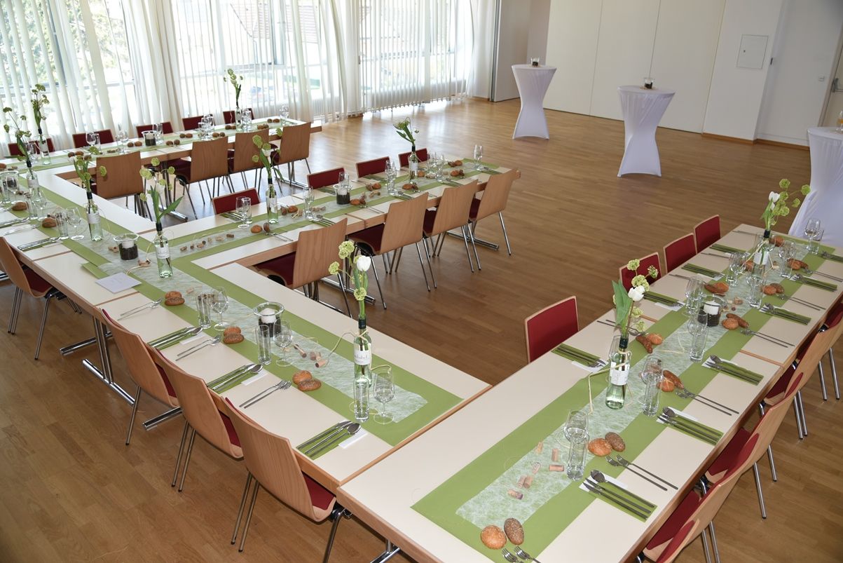 Ein festlich gedeckter Saal mit U-förmig angeordneten Tischen, grünen Tischläufern und Blumendekoration.