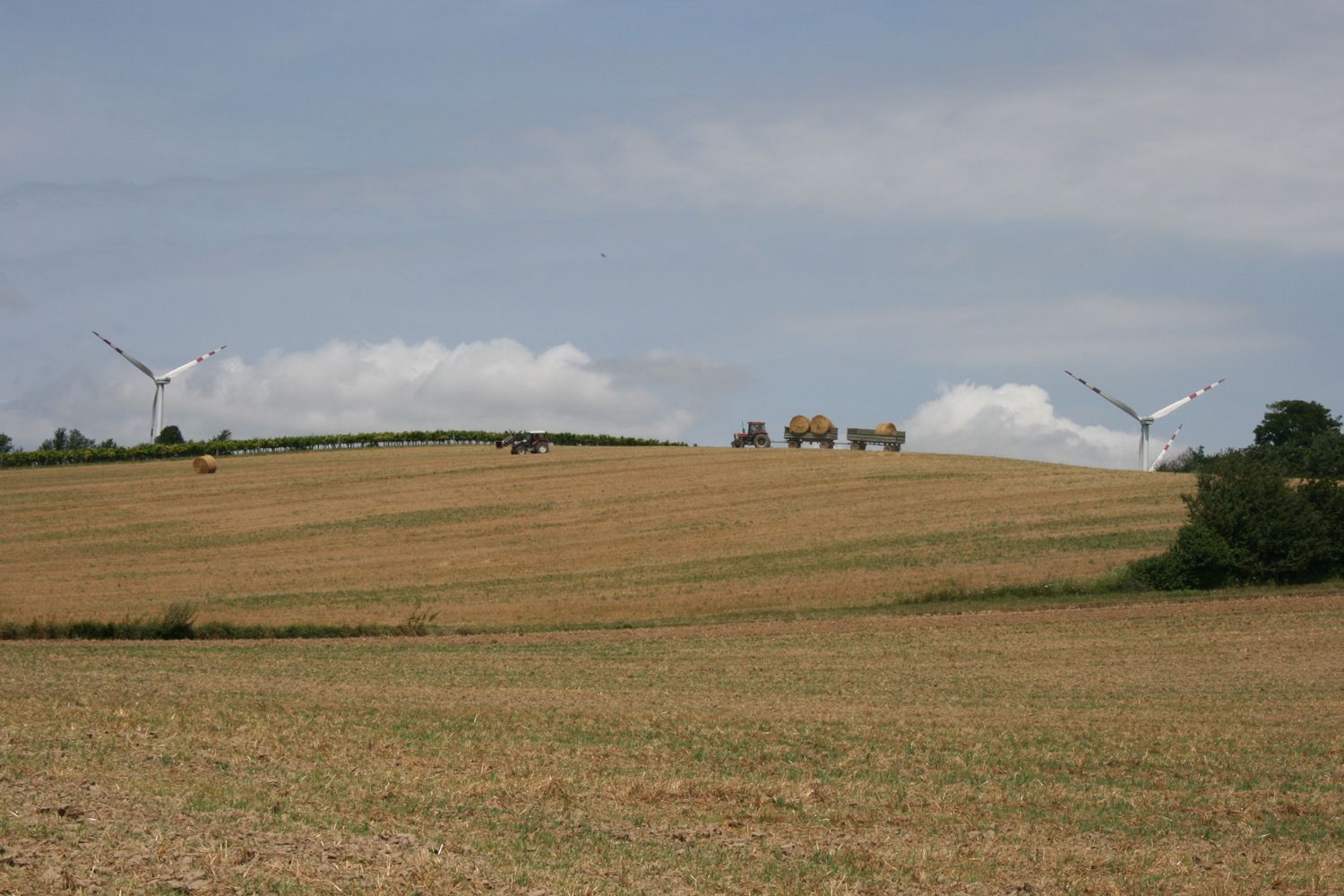 Landschaft mit Windrädern, Traktor und Heuballen auf einem Feld.