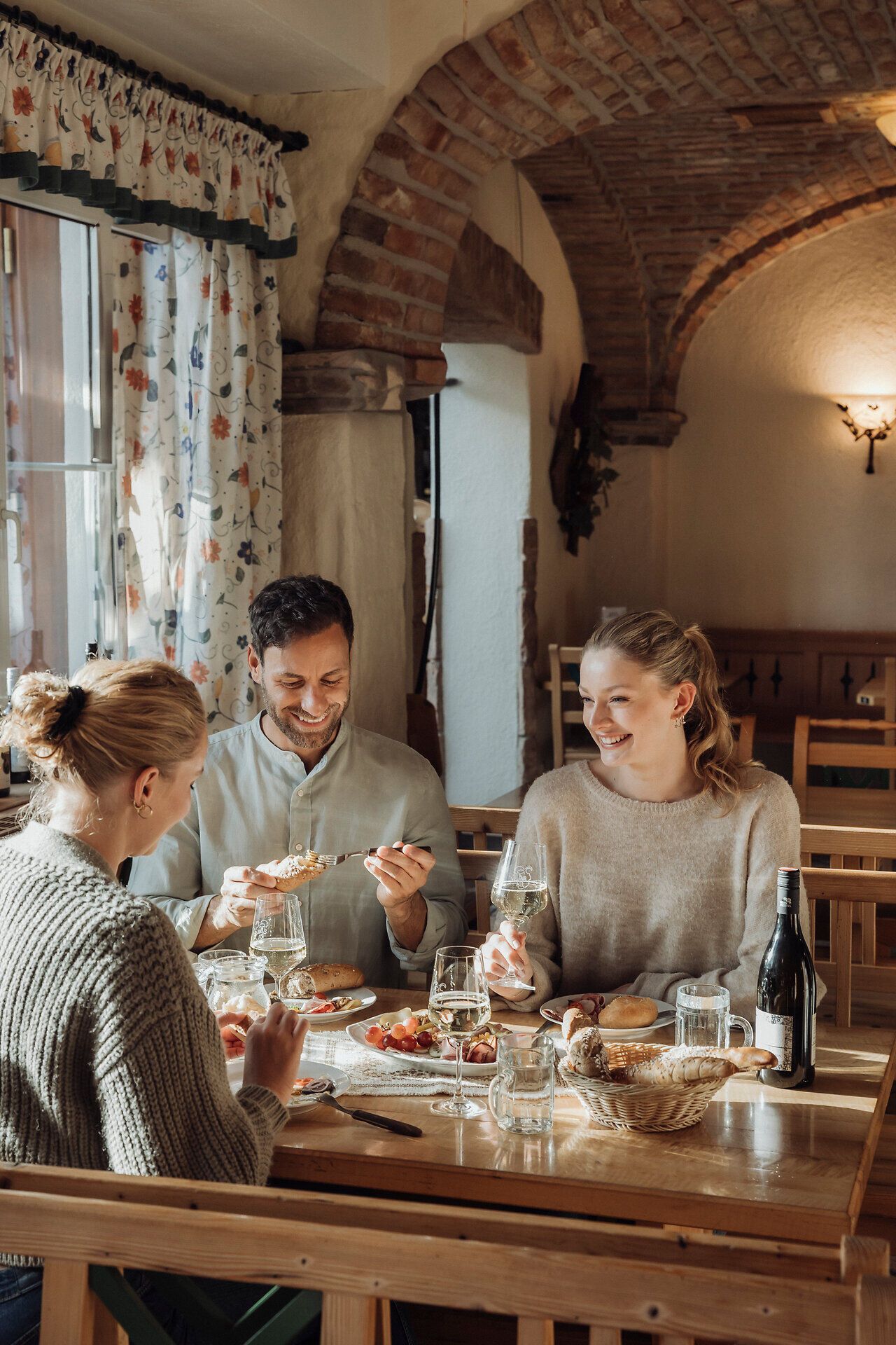 In einem einladenden Heurigenlokal genießen drei Freunde eine köstliche Brettljause, umgeben von rustikalem Charme und der warmen Atmosphäre des Weinviertels. Die Tische sind liebevoll gedeckt, während die Sonne sanft durch die Fenster strahlt und die Geselligkeit fördert.