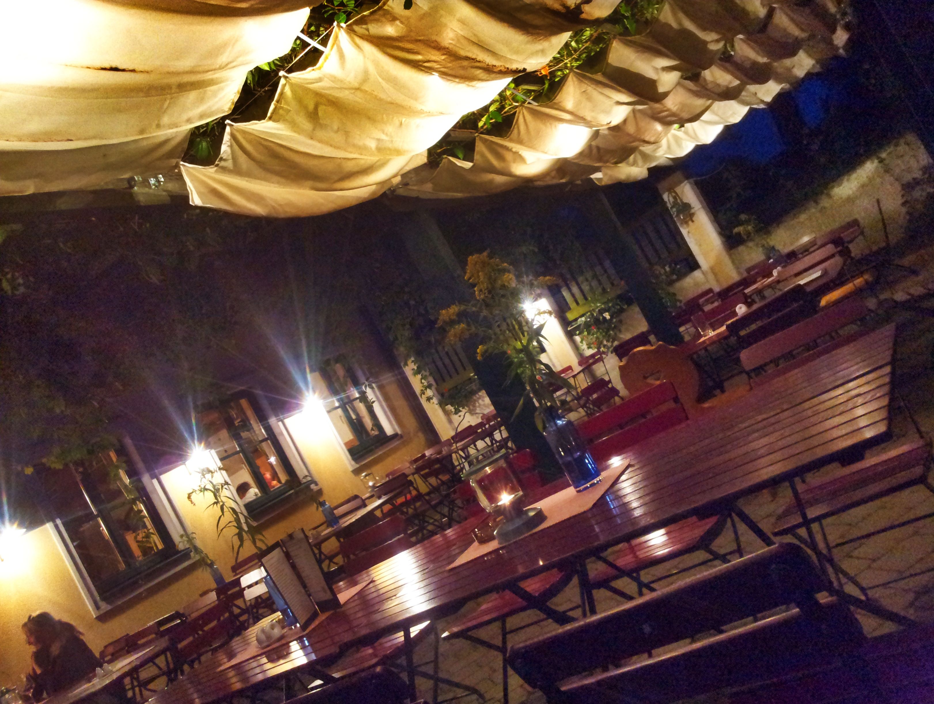 Beleuchtete Terrasse mit Holztischen und Stühlen, überdacht mit Stoffbahnen, bei Nacht.