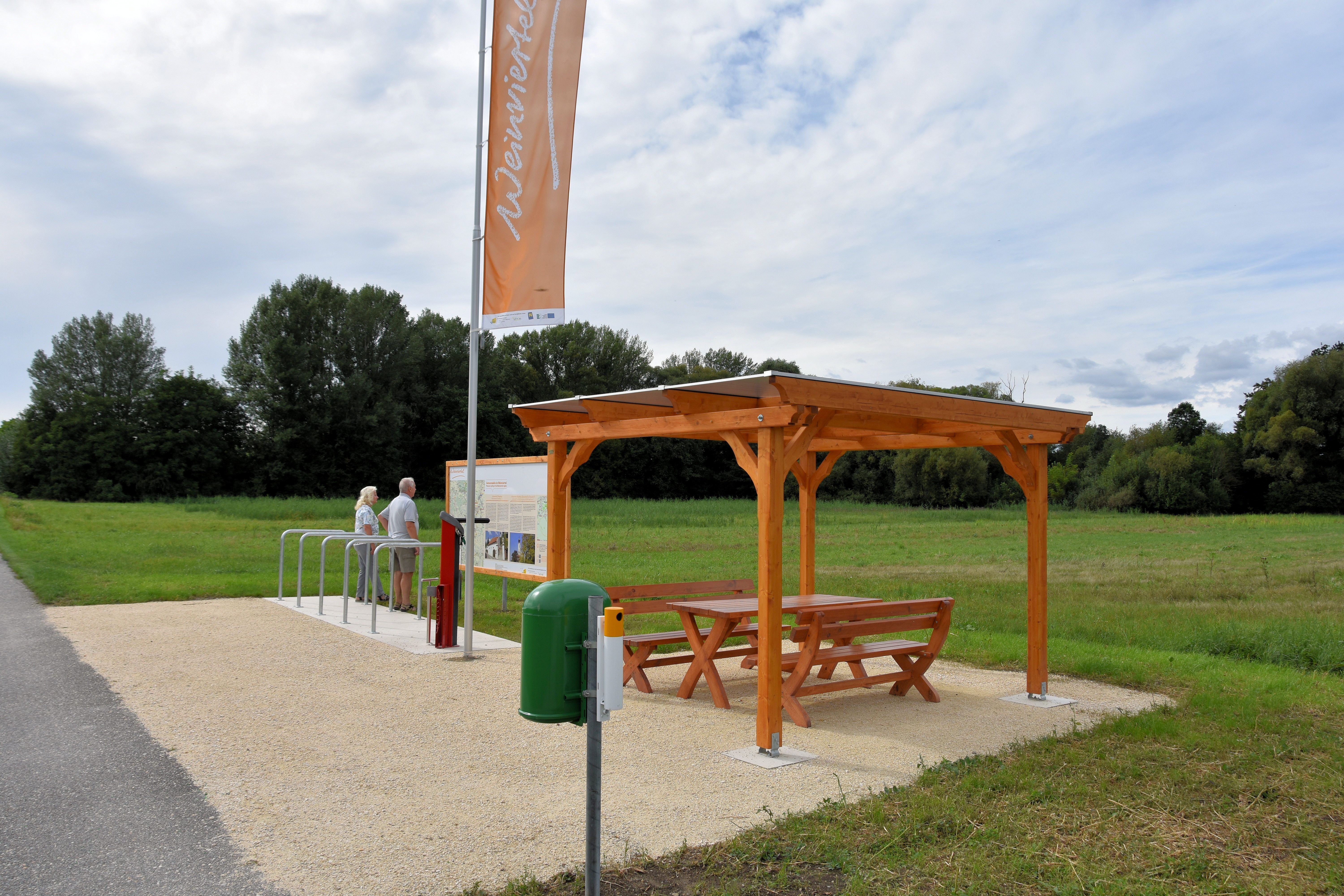 Ein Rastplatz mit Holzdach und Sitzbank neben einem Fahrradständer auf einem Feldweg.