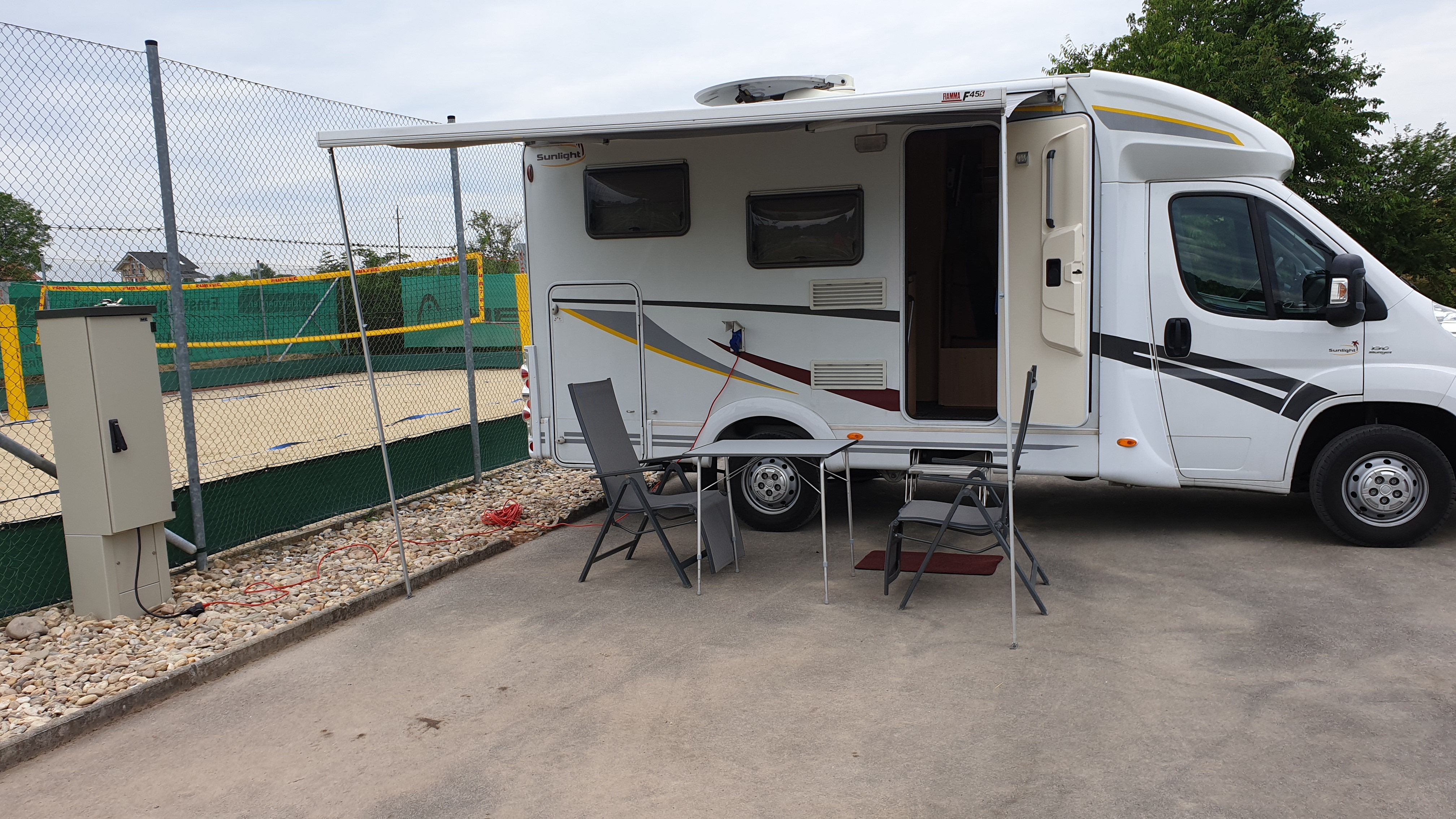 Wohnmobil mit ausgefahrener Markise und Campingmöbeln auf einem Stellplatz neben einem Volleyballfeld.