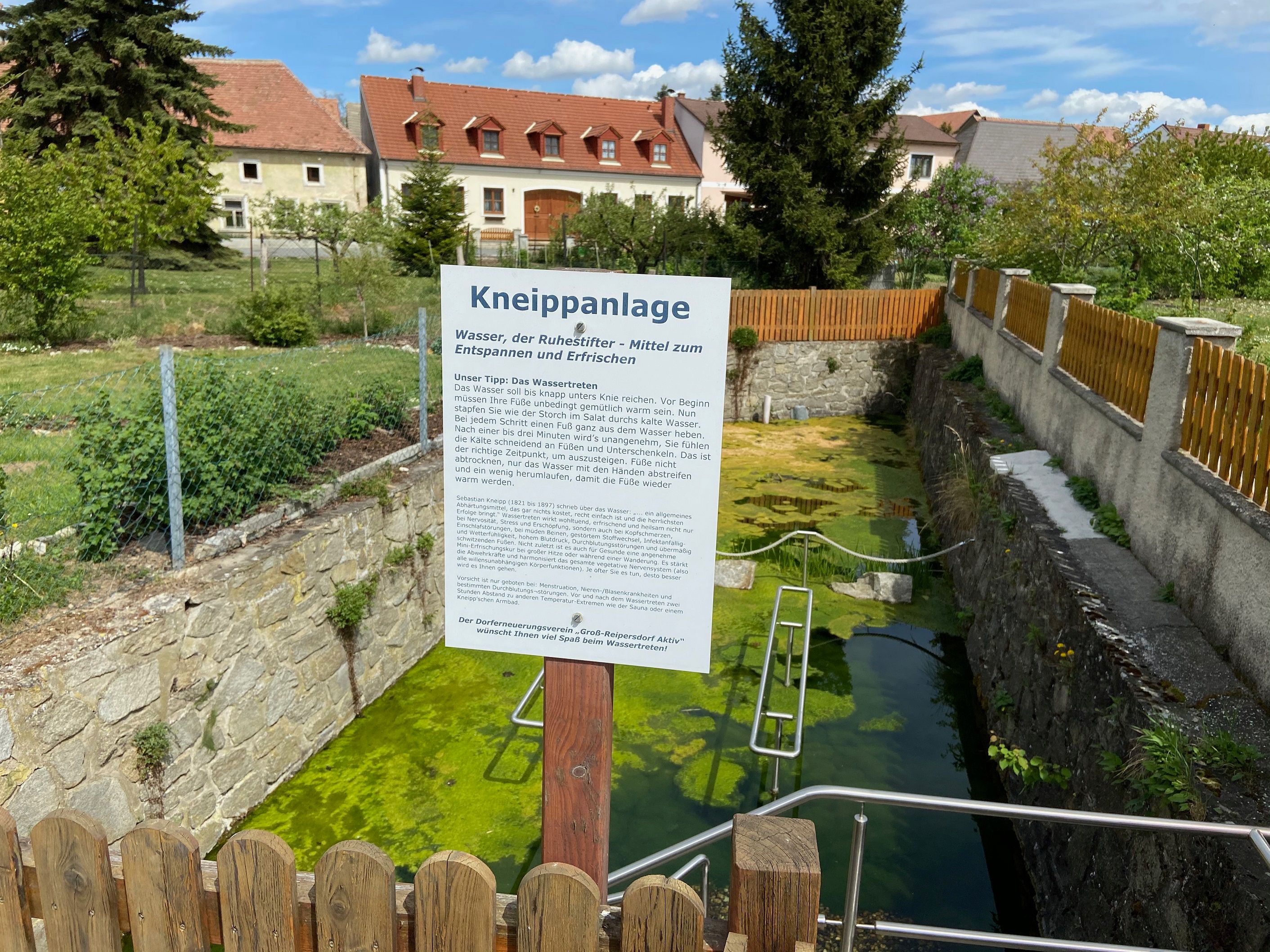 Kneippovo zařízení s informační tabulí a nádrží na vodu, obklopené plotem a domy v pozadí.