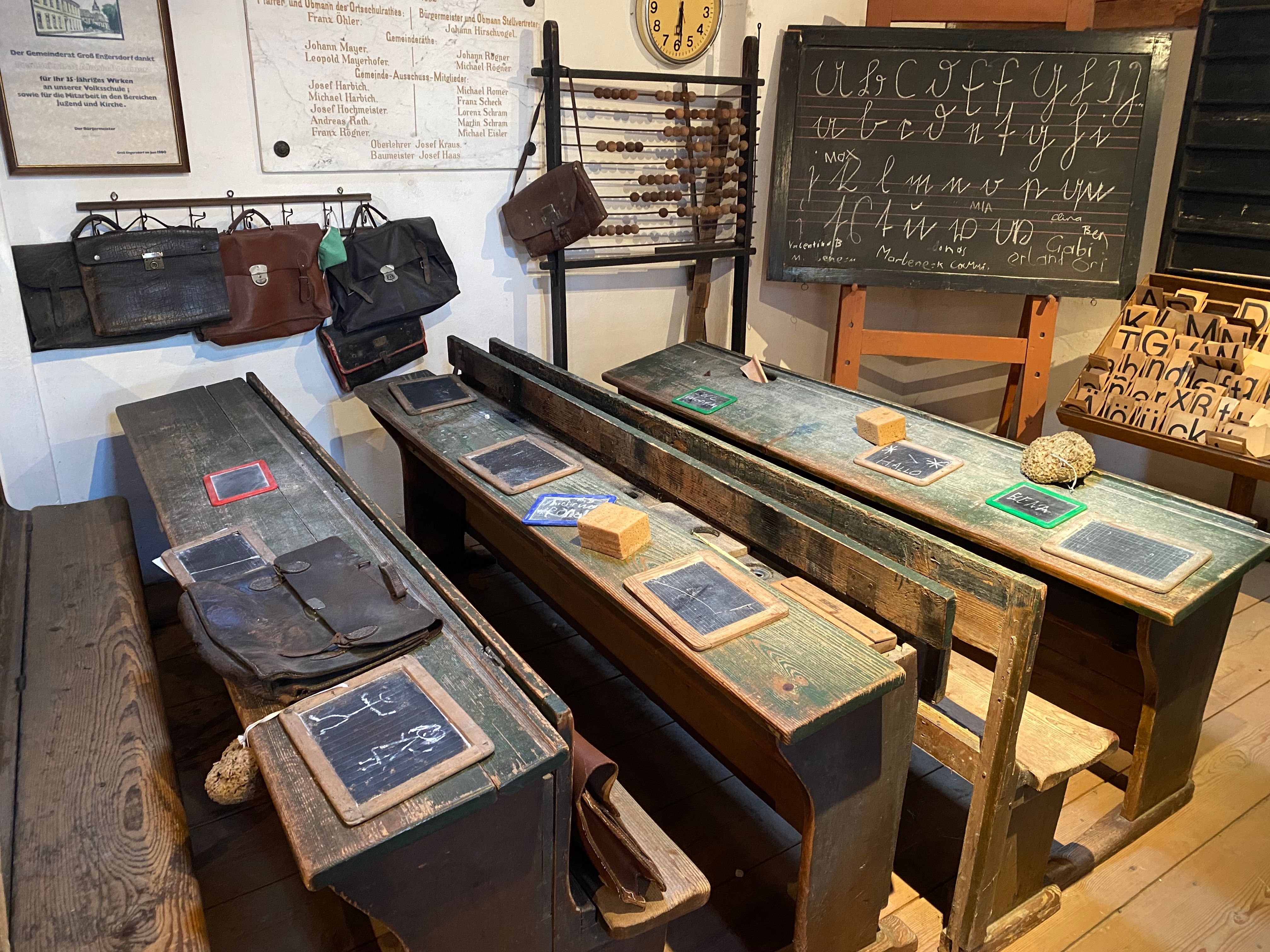 Historisches Klassenzimmer mit alten Holzbänken, Schiefertafeln und einer Tafel mit Schreibübungen.