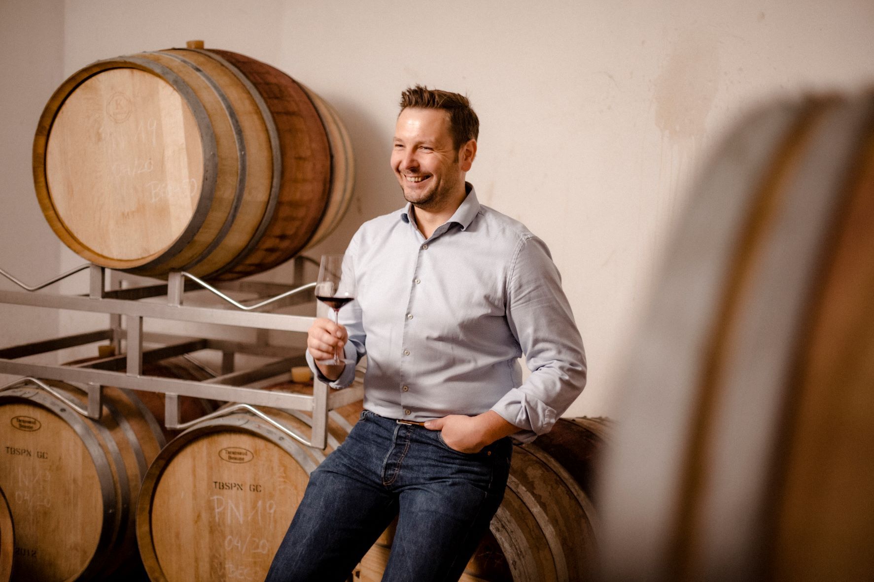 Ein Mann lehnt lächelnd an Weinfässern und hält ein Glas Rotwein in der Hand.