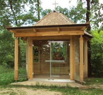 Kleine Holzkapelle mit Glasfront und Kreuz auf dem Dach, umgeben von Bäumen.