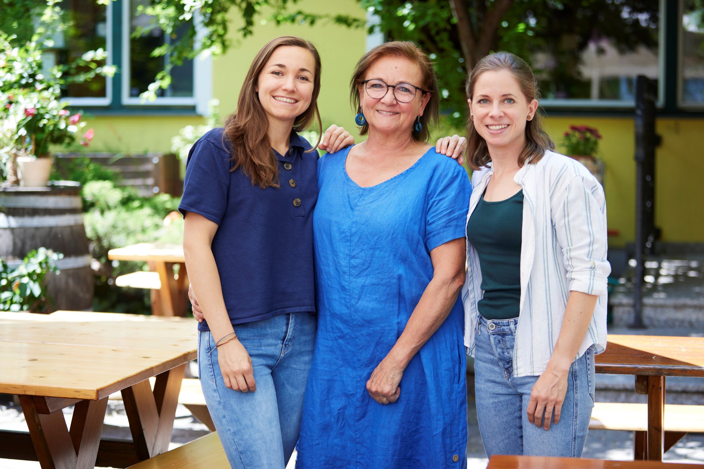 Drei Frauen stehen lächelnd in einem Garten mit Holztischen und Pflanzen.