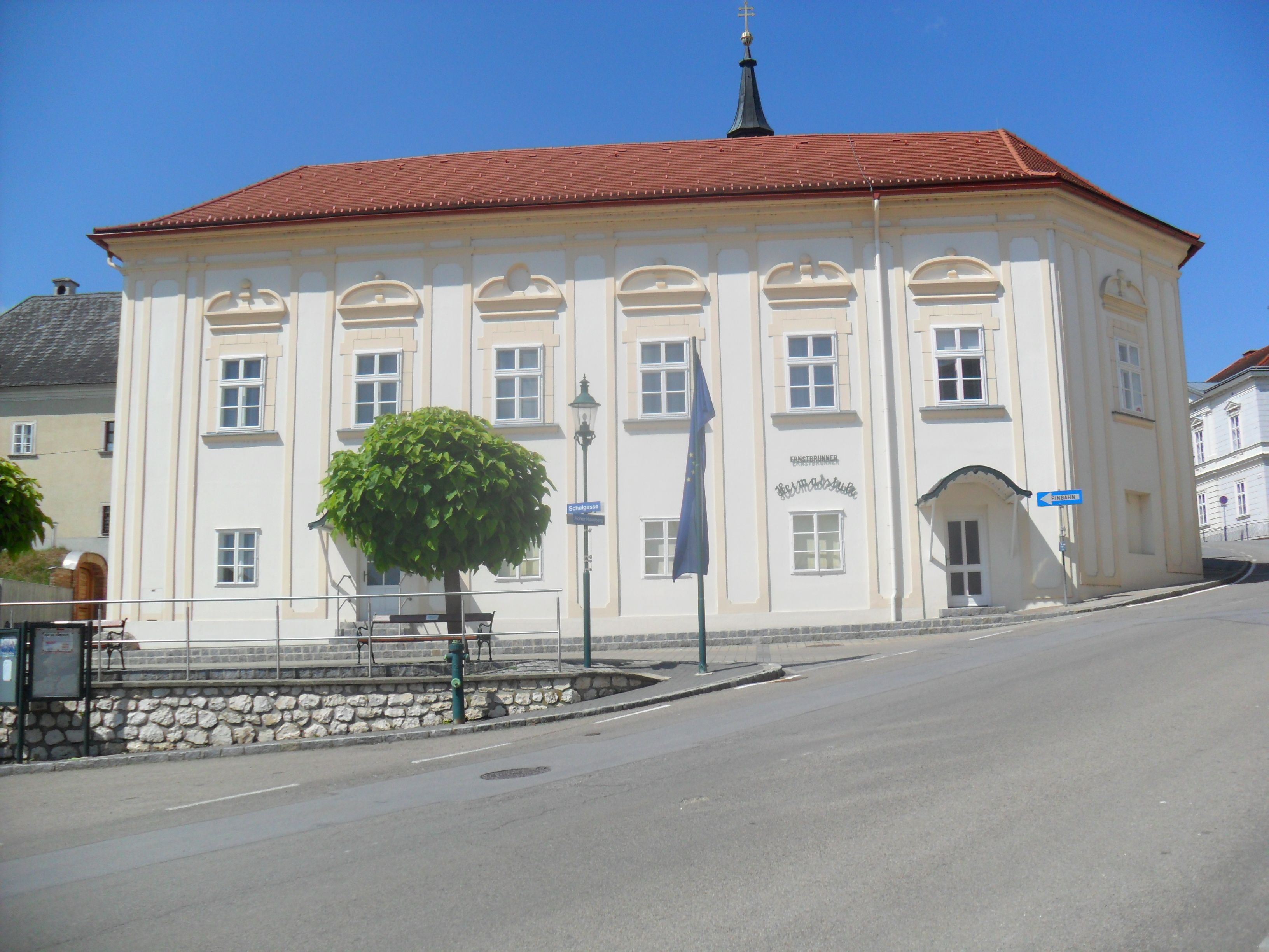 Historisches Gebäude mit rotem Dach und weißen Wänden, umgeben von Bäumen und einer Straße.