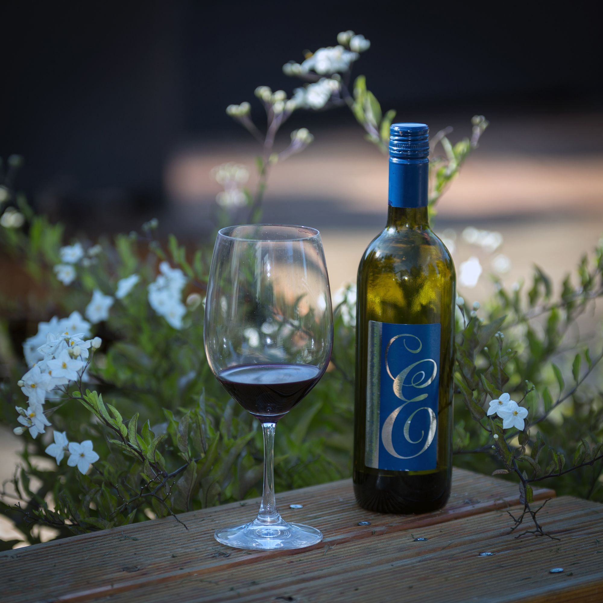 Weinflasche und Glas auf Holztisch mit Blumen im Hintergrund.