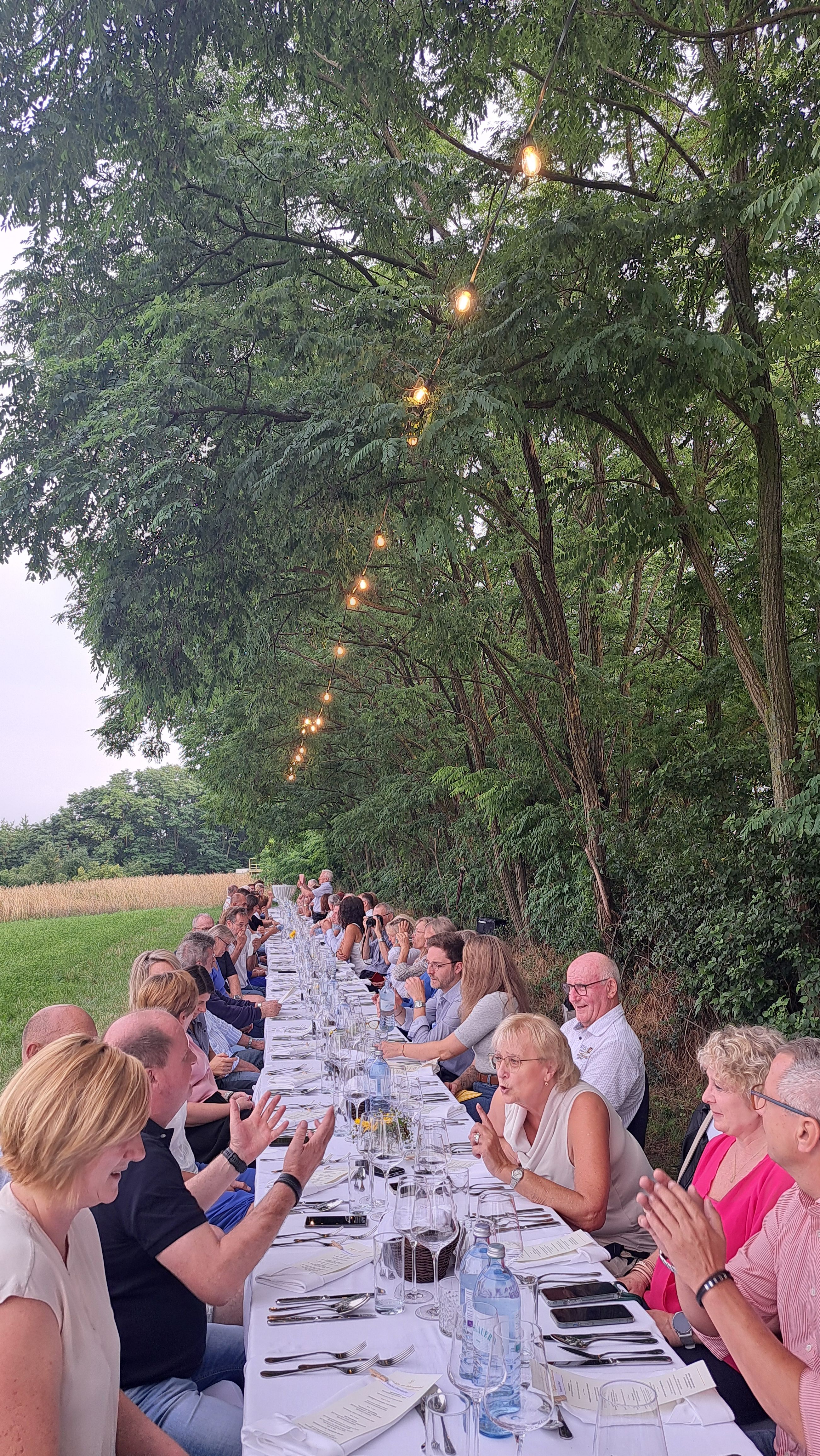 Eine lange Tafel im Freien mit vielen Menschen, die unter Bäumen speisen. Lichterketten hängen über dem Tisch.