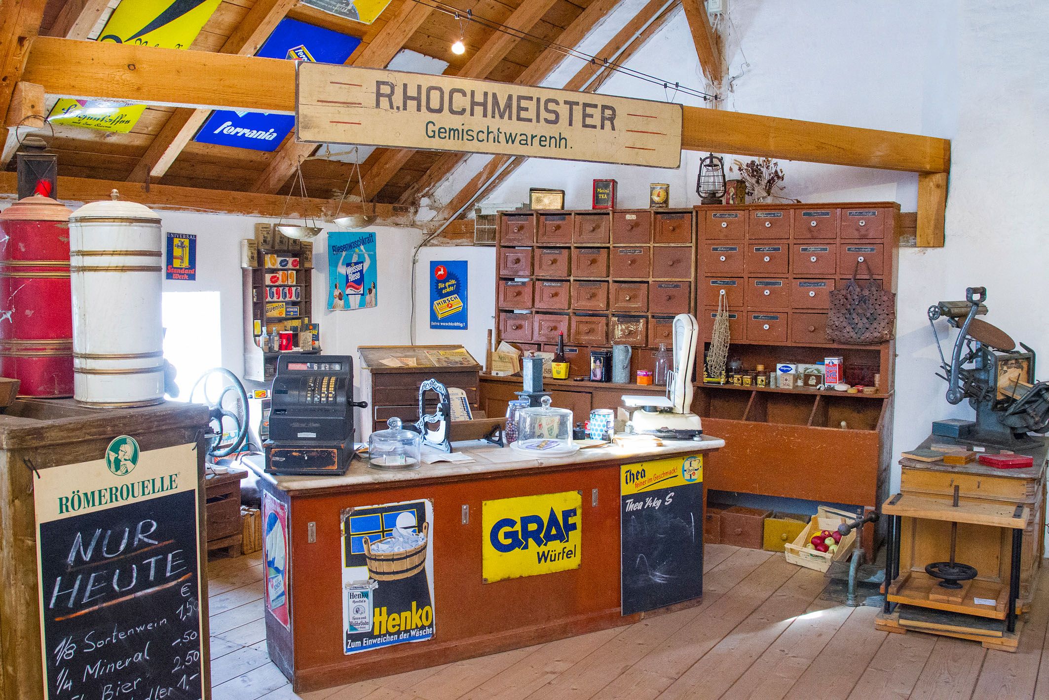 Innenansicht eines alten Gemischtwarenladens mit Holzmöbeln, einer Registrierkasse und Werbeschildern an den Wänden.