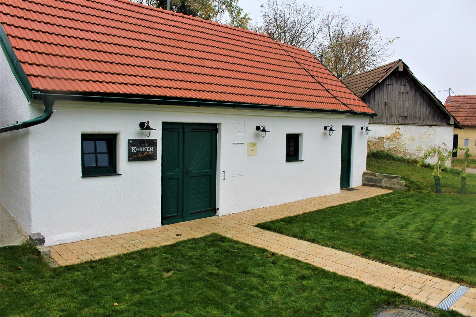 Biela budova s červenou strechou a zelenými dverami, obklopená trávnikom.