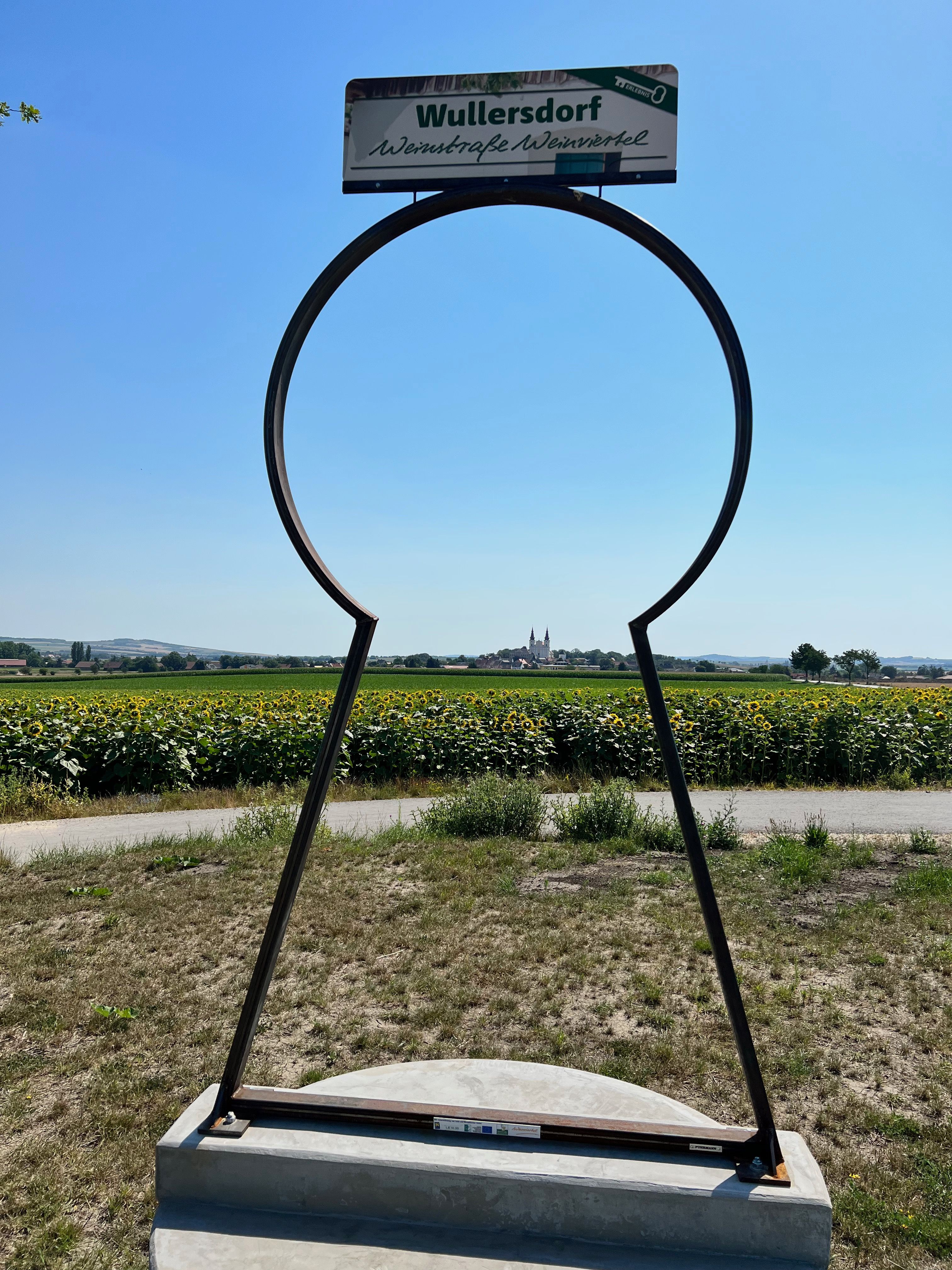 Metallrahmen in Form eines Schlüssellochs mit Blick auf Wullersdorf und Sonnenblumenfeld.