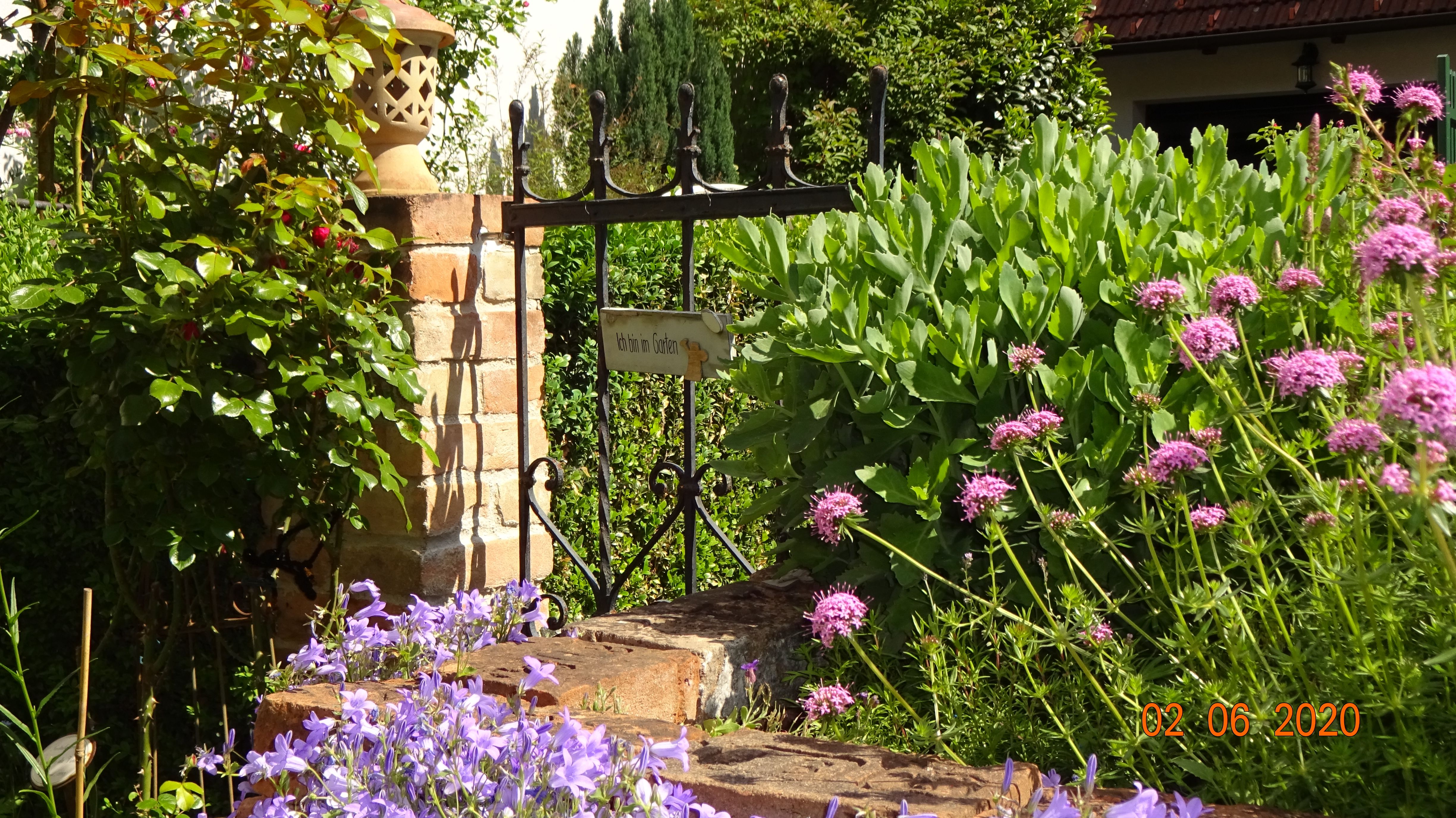 Ein Garten mit einem kleinen Tor und einem Schild mit der Aufschrift 'Ich bin im Garten'. Umgeben von blühenden Pflanzen und einem Ziegelsteinpfosten.