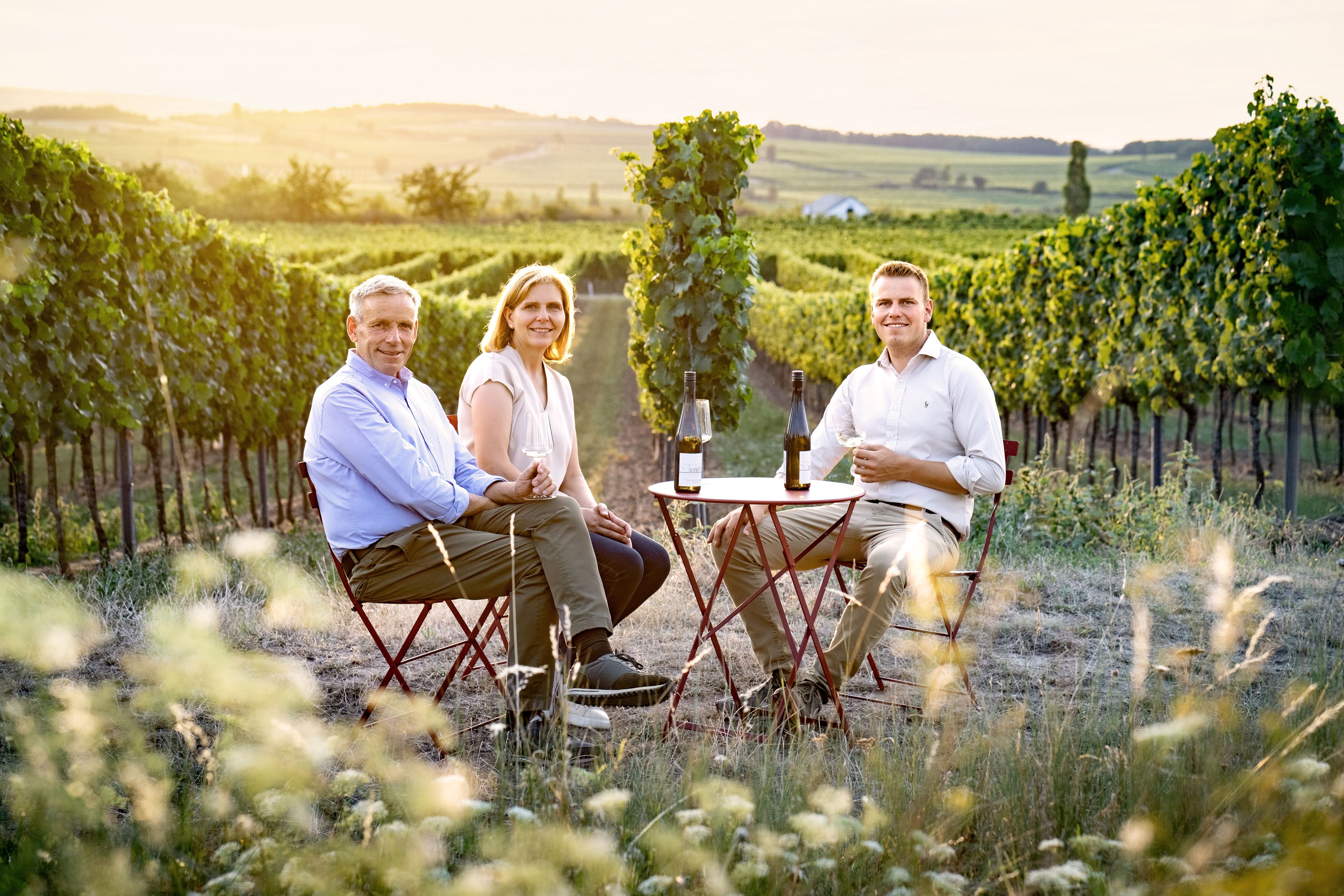 Drei Personen sitzen an einem Tisch im Weinberg mit Weingläsern und Flaschen.