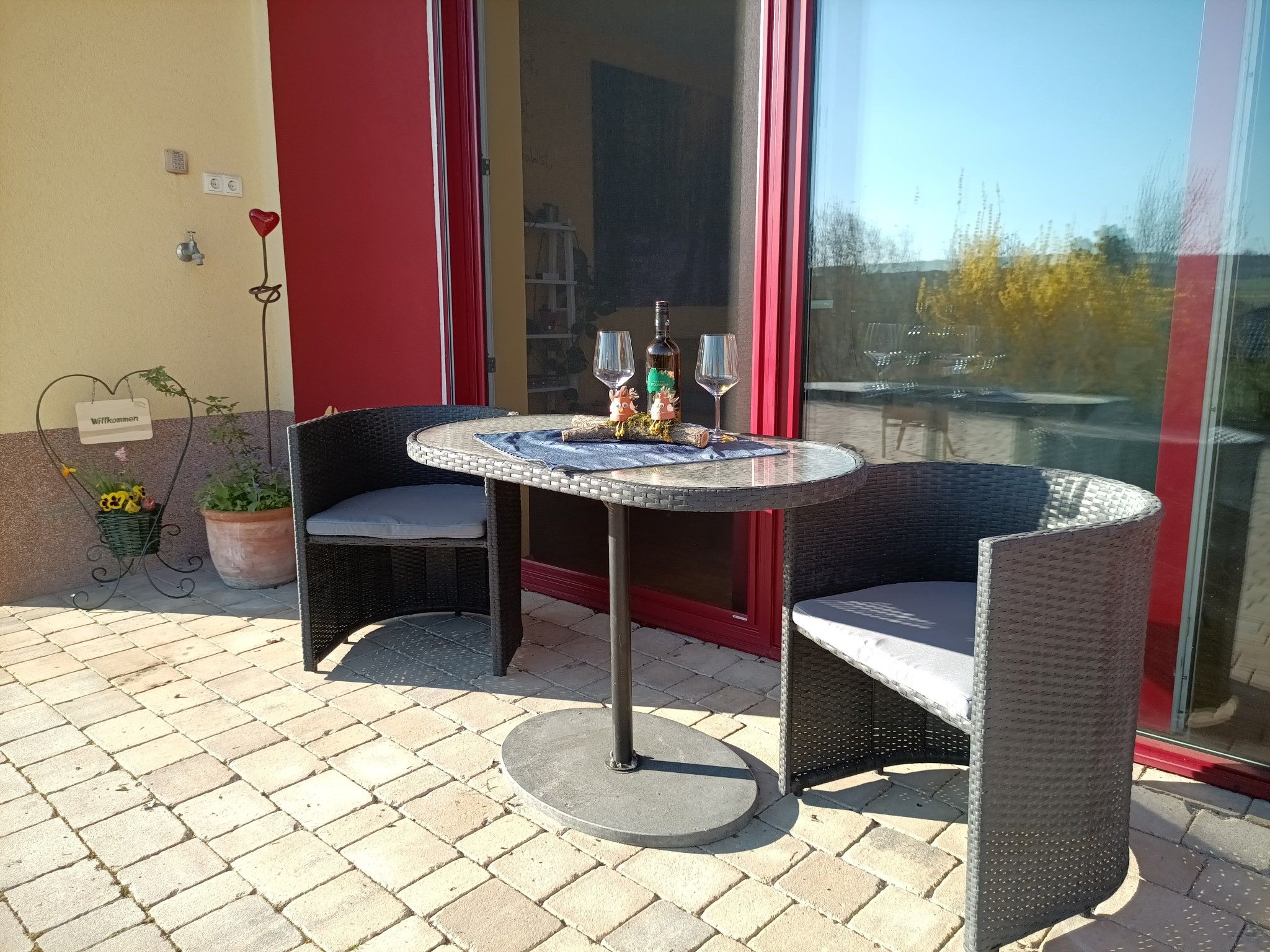 Terrasse mit Tisch, zwei Stühlen, Weinflasche und Gläsern vor einer roten Tür.