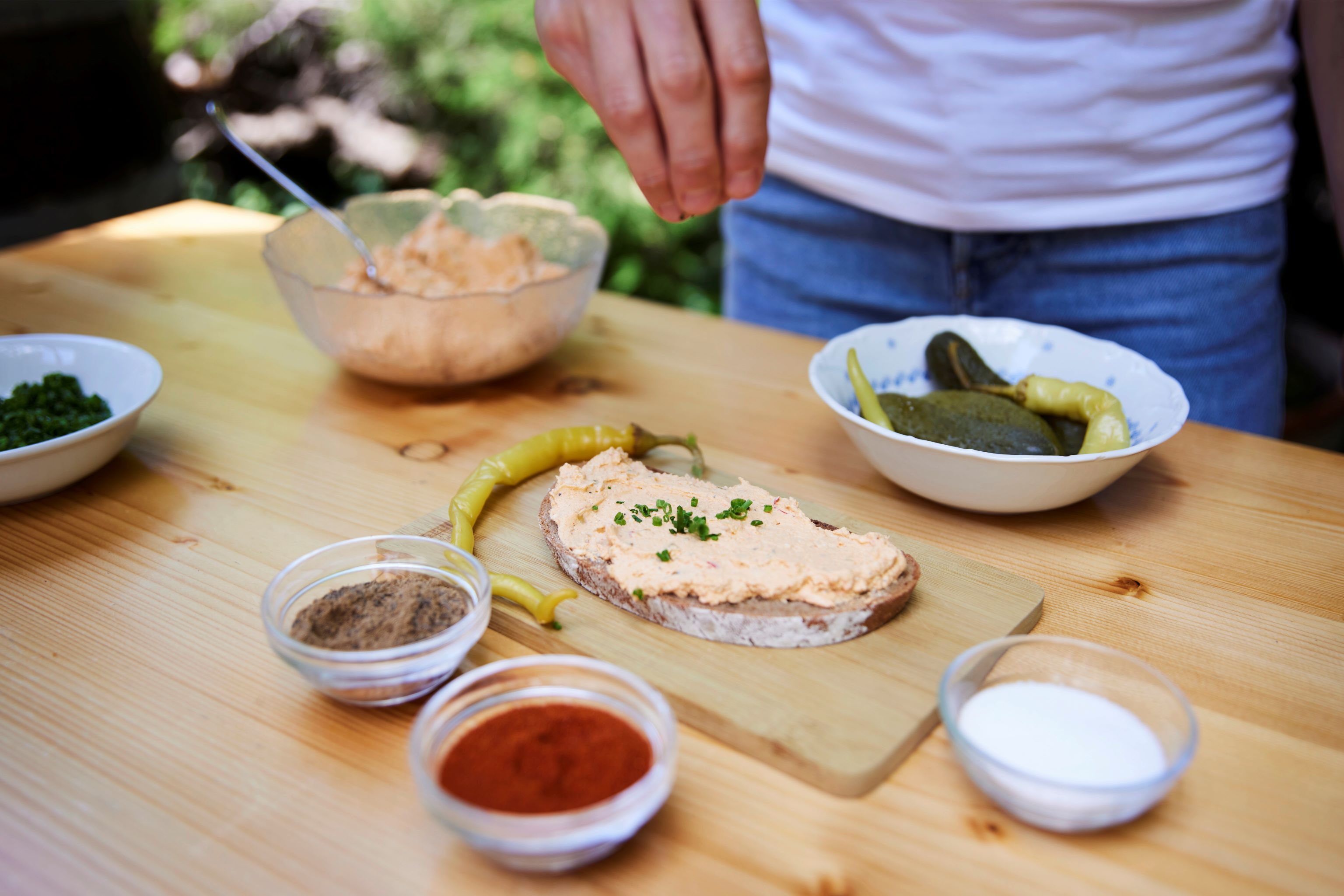Ein Liptauerbrot auf einem Holzbrett, umgeben von Gewürzen und eingelegtem Gemüse.