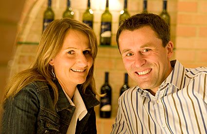 A smiling couple in a wine cellar with bottles of wine in the background.