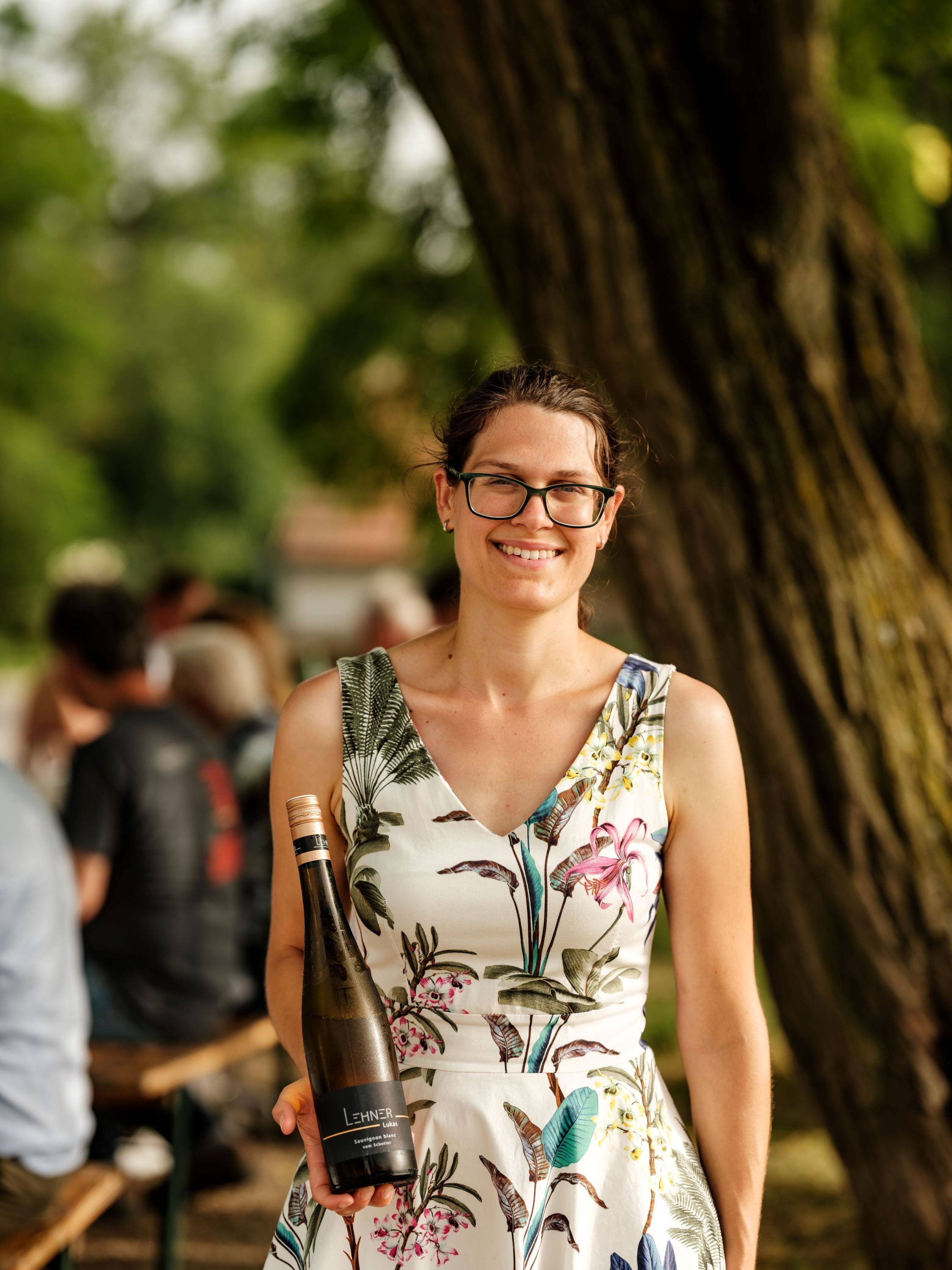 Woman with glasses holding a wine bottle outdoors.