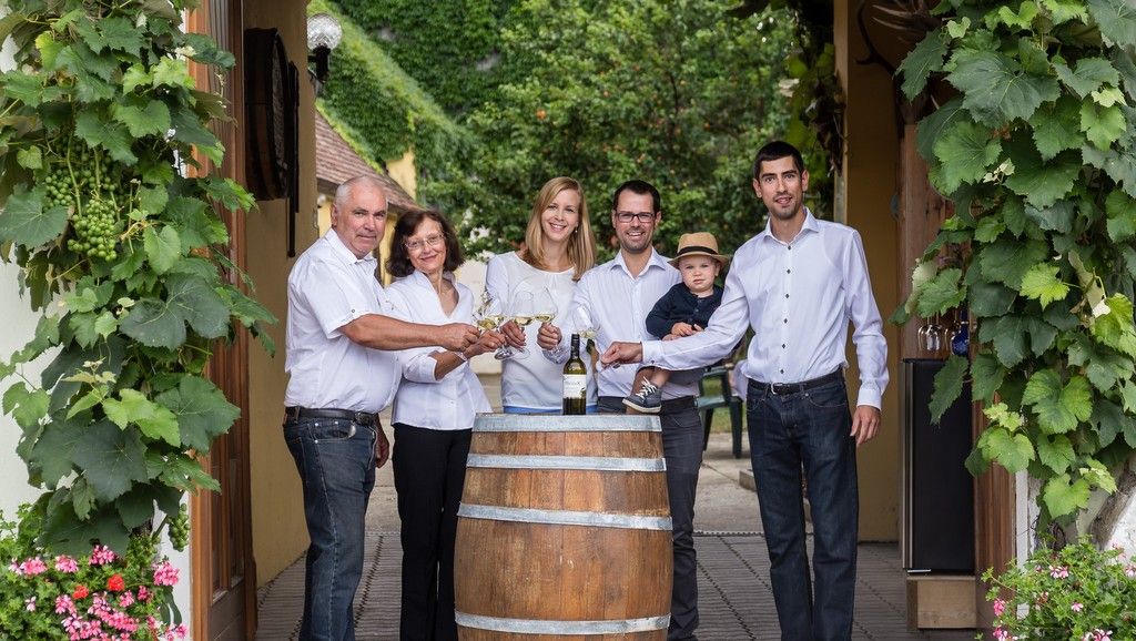 Eine Familie steht um ein Weinfass, umgeben von Weinreben, und stößt mit Weingläsern an.