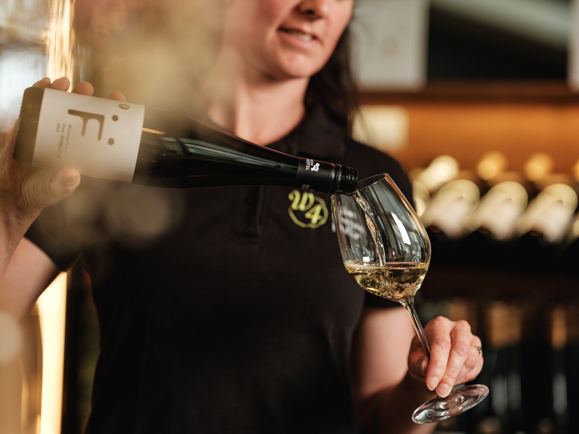 In einer einladenden Vinothek wird ein edler Wein in ein elegantes Glas gegossen. Die warmen Farben des Weins spiegeln die goldene Herbstsonne wider und laden dazu ein, die Aromen der Region zu entdecken. Ein perfekter Moment, um die kulinarischen Köstlichkeiten des Weinviertels zu genießen.