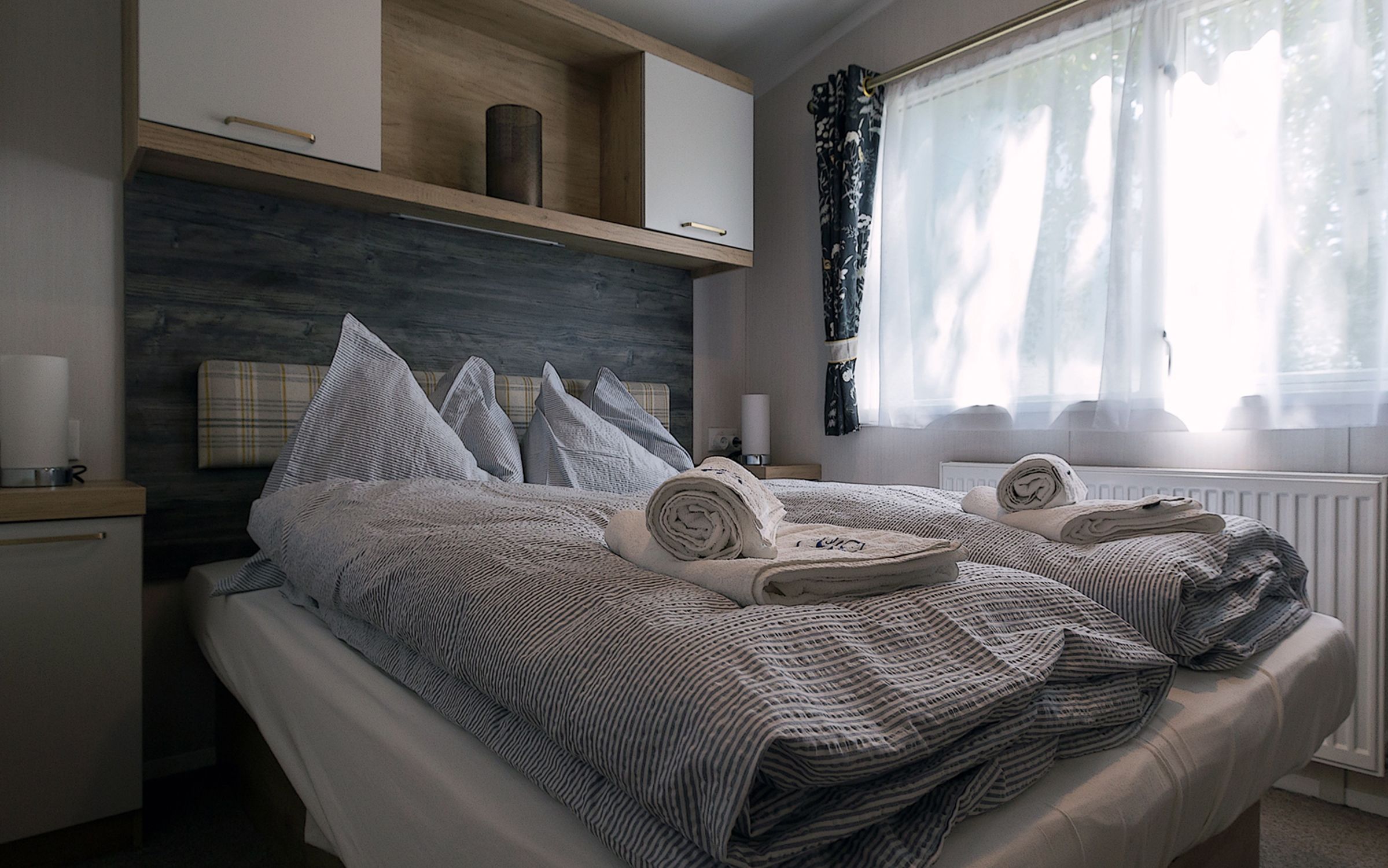Ein gemütliches Schlafzimmer mit einem Doppelbett, grauer Bettwäsche und gerollten Handtüchern. Ein Fenster mit Vorhängen lässt Tageslicht herein.