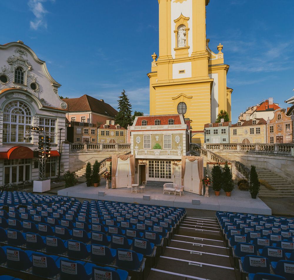 Ein malerisches Freilufttheater, umgeben von historischen Gebäuden, lädt Besucher zu unvergesslichen Aufführungen ein. Die bunten Stühle sind bereit, die Zuschauer zu empfangen, während die strahlende Sonne den Himmel erleuchtet und eine festliche Stimmung verbreitet.