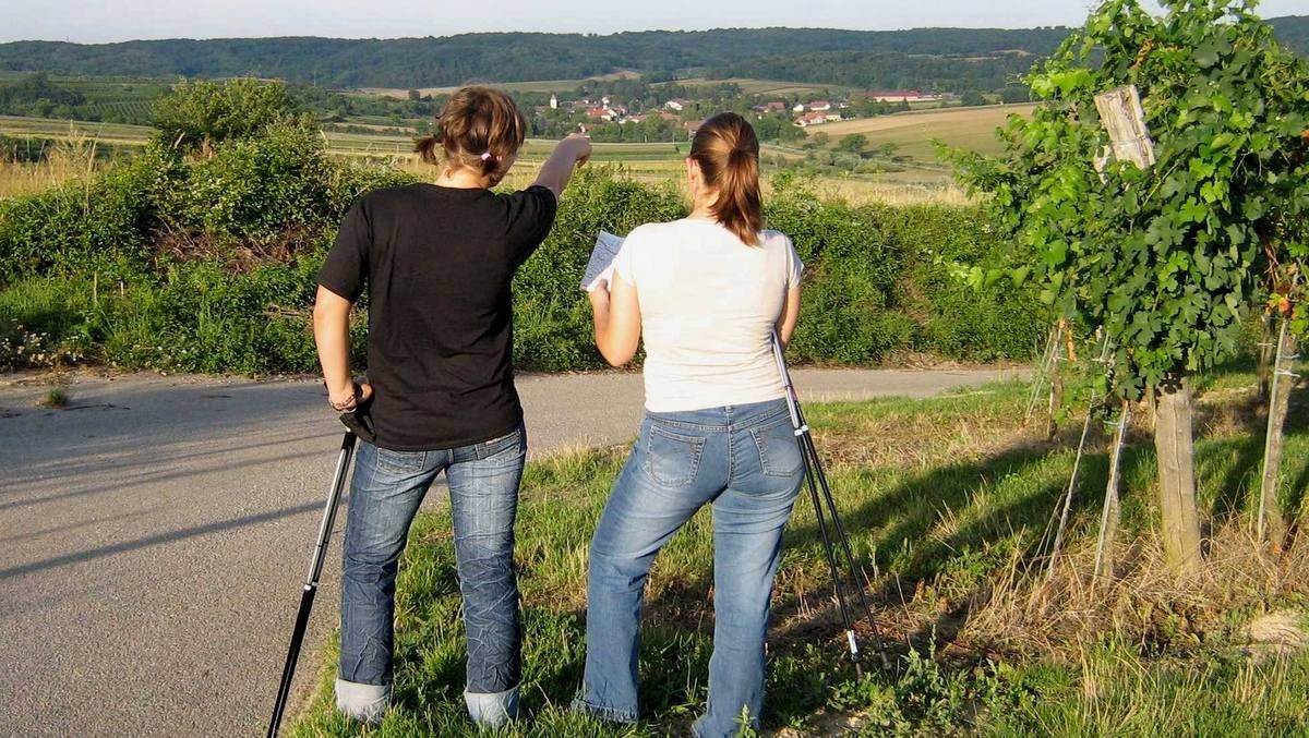 Zwei Frauen mit Wanderstöcken stehen auf einem Weg und schauen in die Ferne, eine zeigt in die Richtung eines Dorfes.