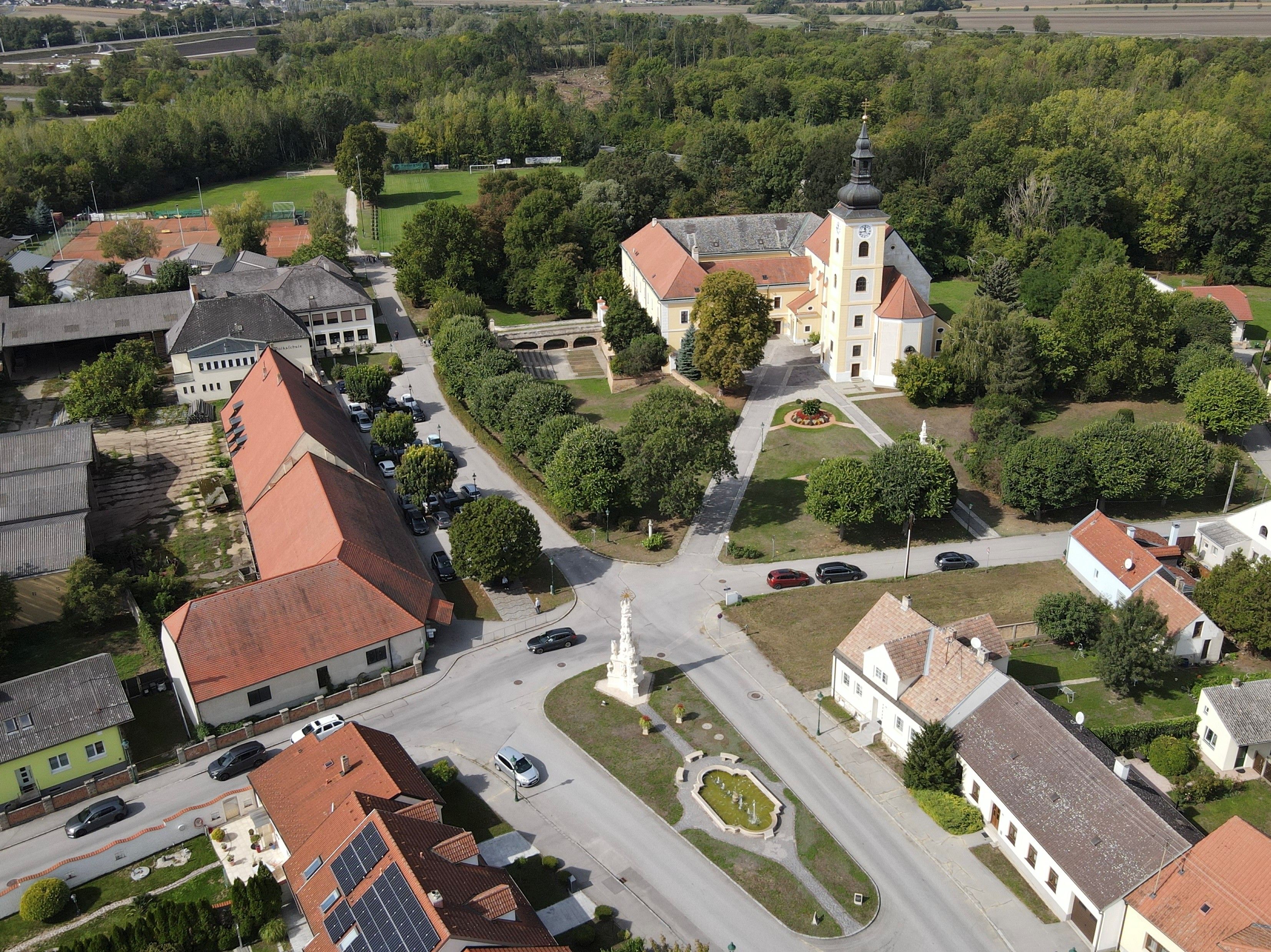 Luftaufnahme von Weikendorf mit Kirche, Dorfplatz, Brunnen und umliegenden Häusern am Rand eines Waldgebiets.