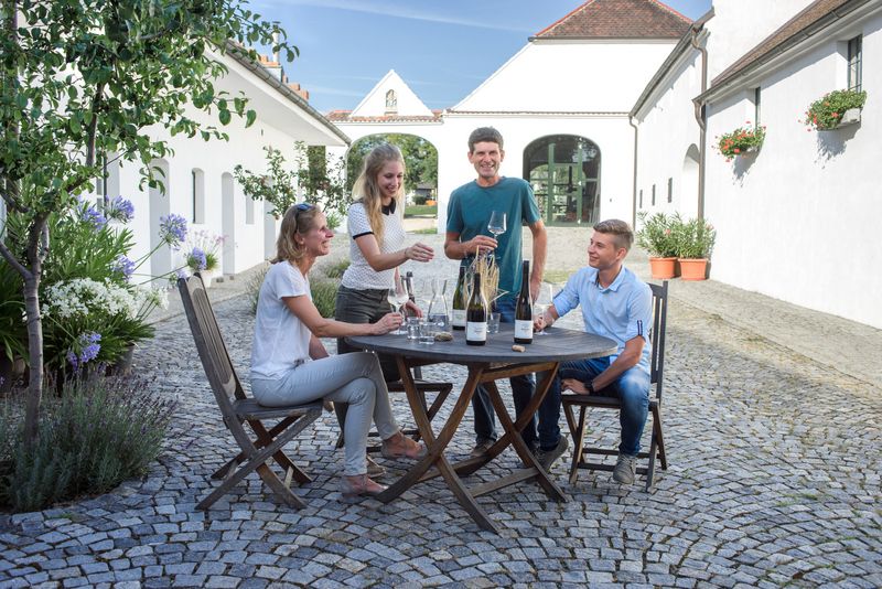 Vier Personen sitzen an einem runden Tisch im Freien und genießen Wein. Sie sind von weißen Gebäuden umgeben.