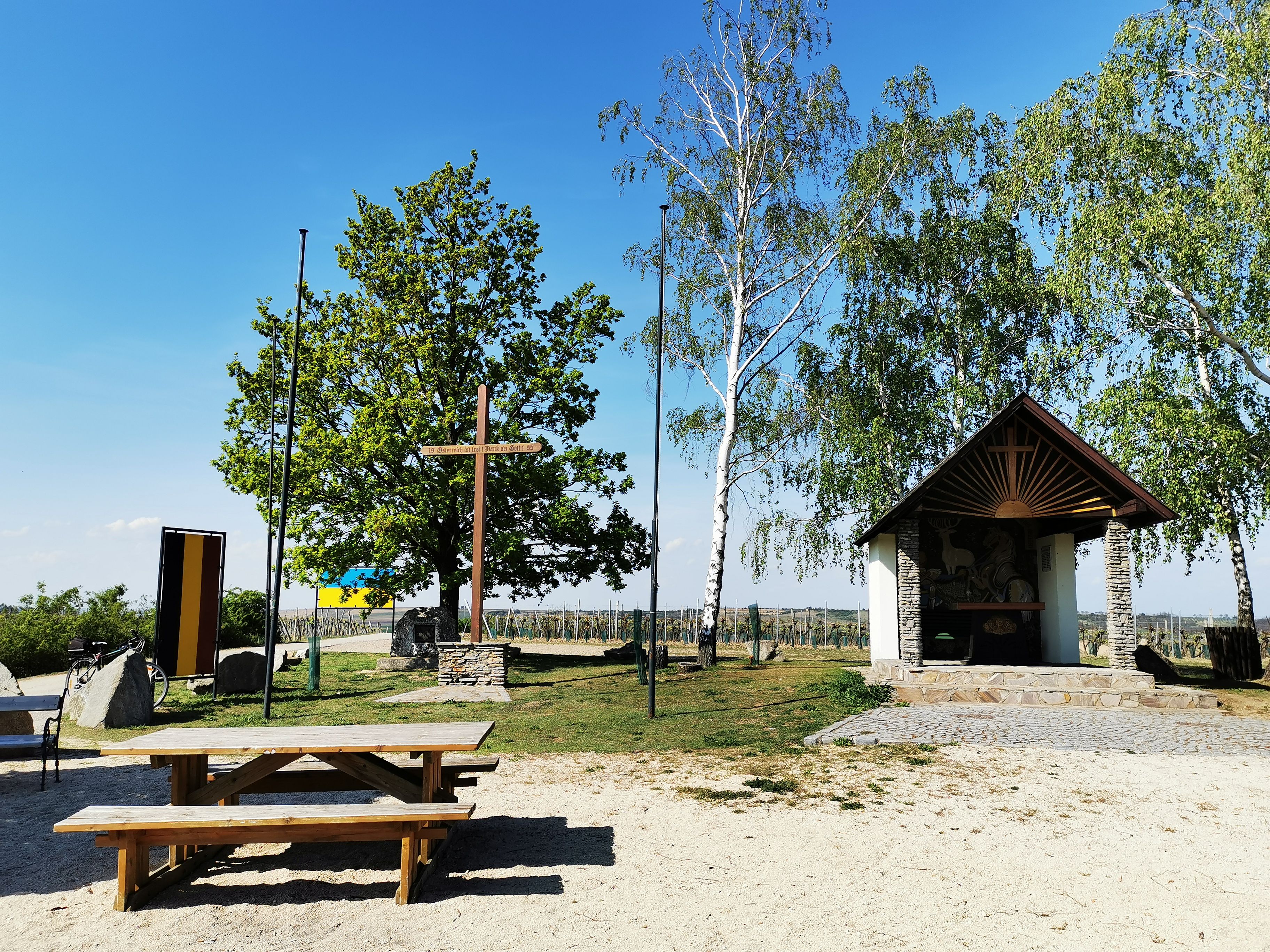 Hubertuskapelle Hadres mit Holzkreuz, Bäumen und Picknicktisch bei sonnigem Wetter.
