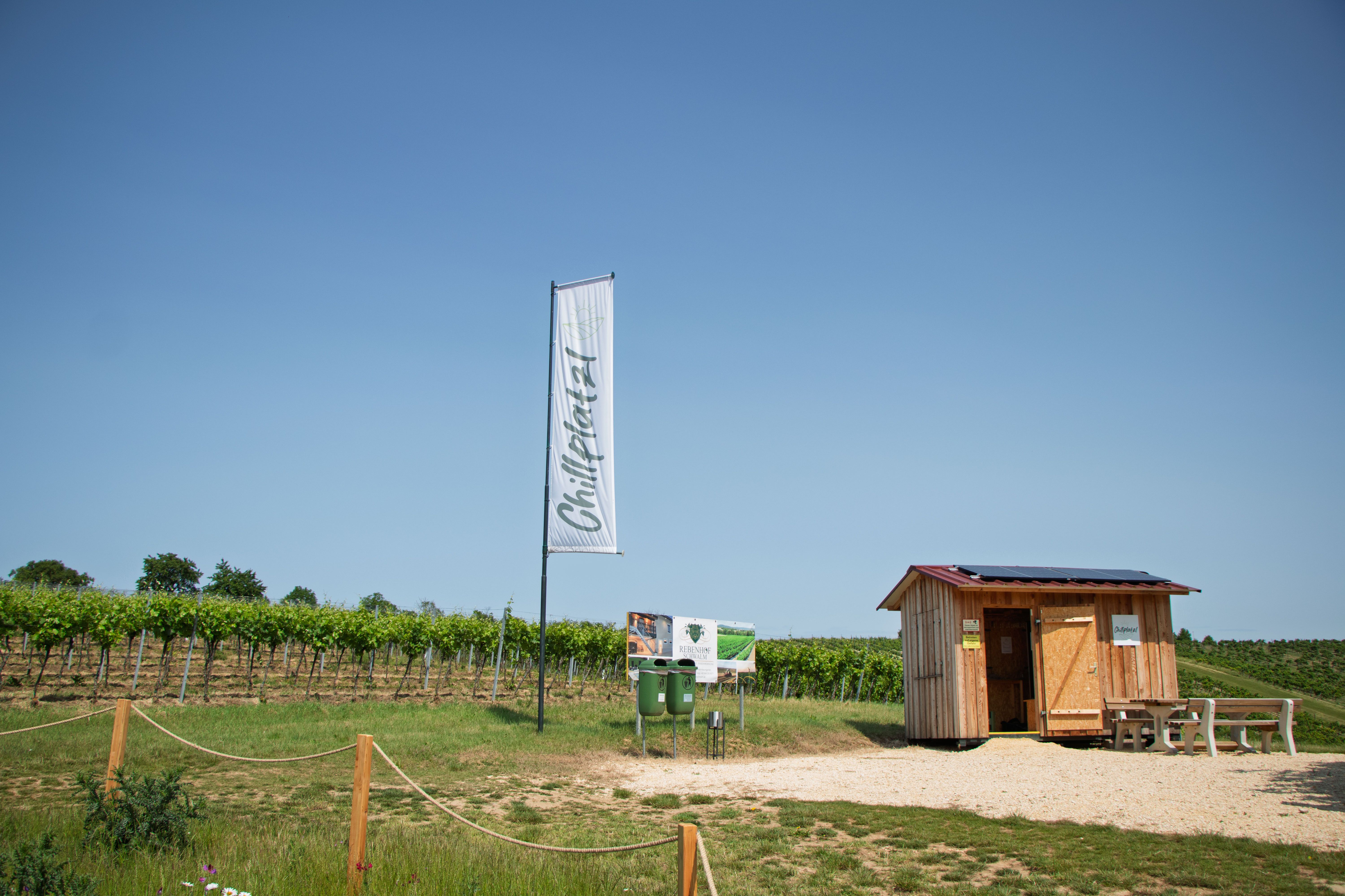 Ein kleiner Holzschuppen neben einem Weinberg mit einem Schild und einer Fahne mit der Aufschrift 'Chillplatz'.
