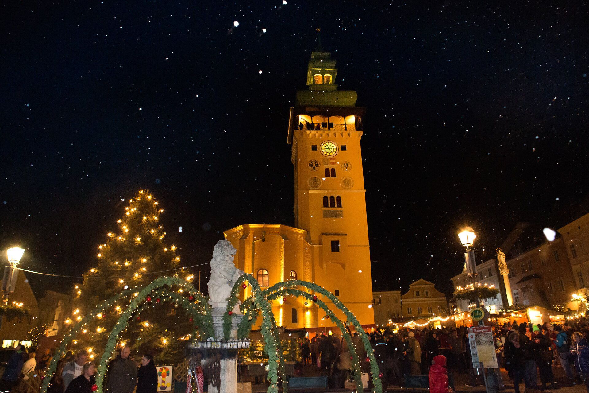 In der festlich beleuchteten Stadt erstrahlt der historische Turm in warmem Licht, während der Duft von Glühwein und frisch gebackenem Gebäck die Luft erfüllt. Umgeben von fröhlichen Menschen und einem prächtigen Weihnachtsbaum, entsteht eine zauberhafte Atmosphäre, die zum Verweilen einlädt.