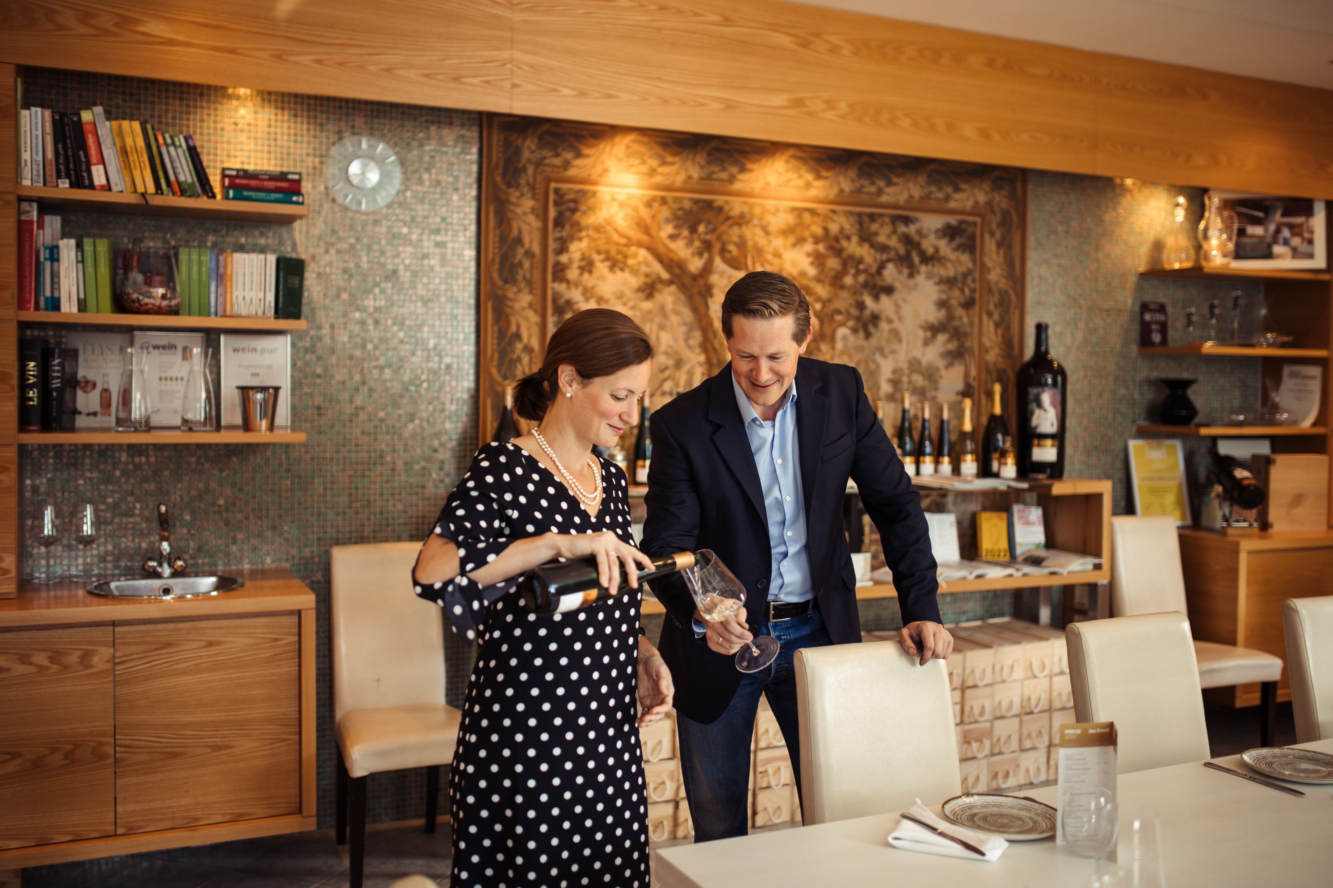 Ein Winzerpaar steht in einem edlen Verkostungsraum, die Frau schenkt dem Mann ein Glas Weißwein ein.