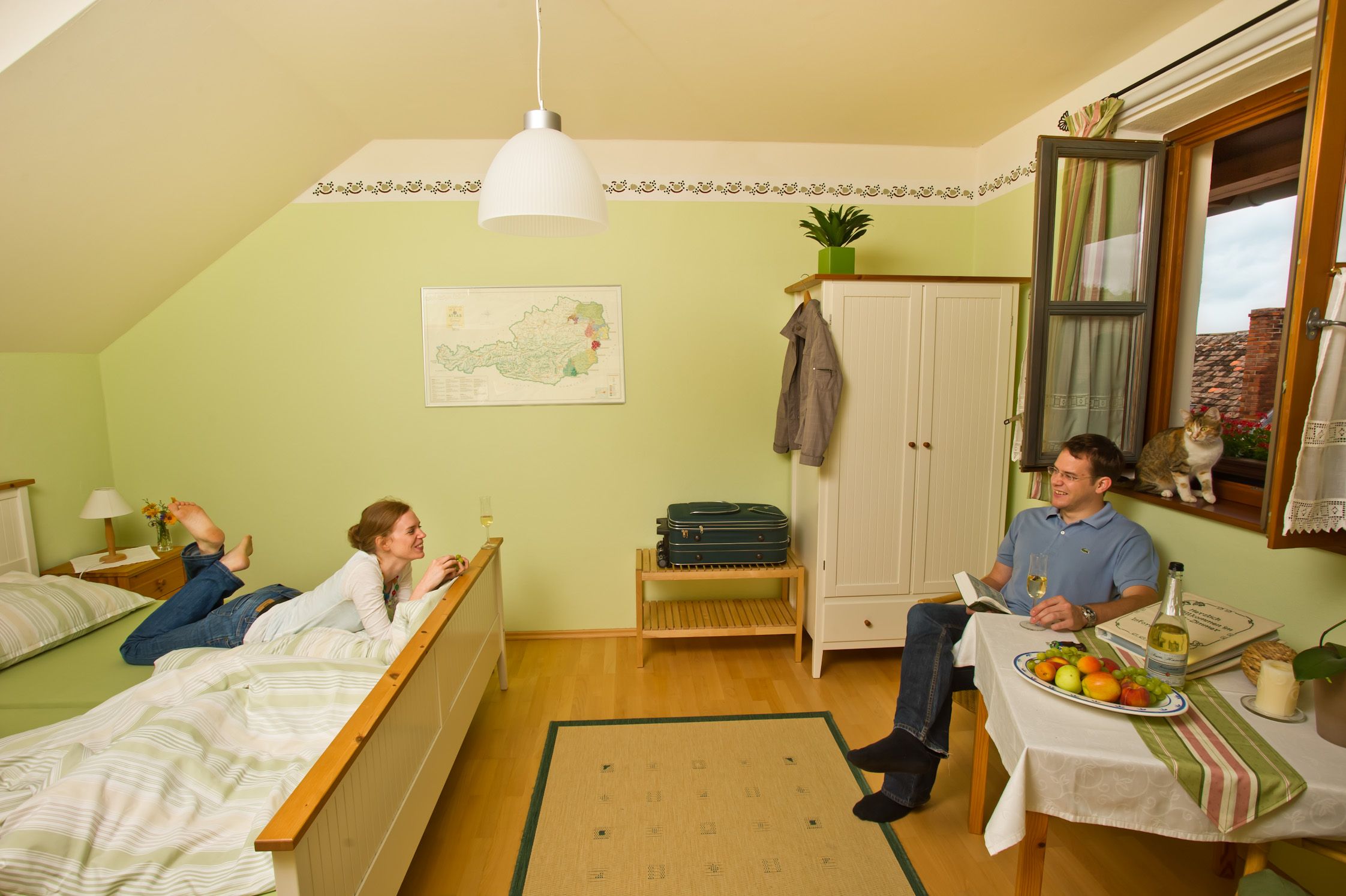 Ein gemütliches Gästezimmer mit einem Paar, das entspannt. Eine Frau liegt auf dem Bett, ein Mann sitzt am Tisch. Eine Katze sitzt am Fenster.