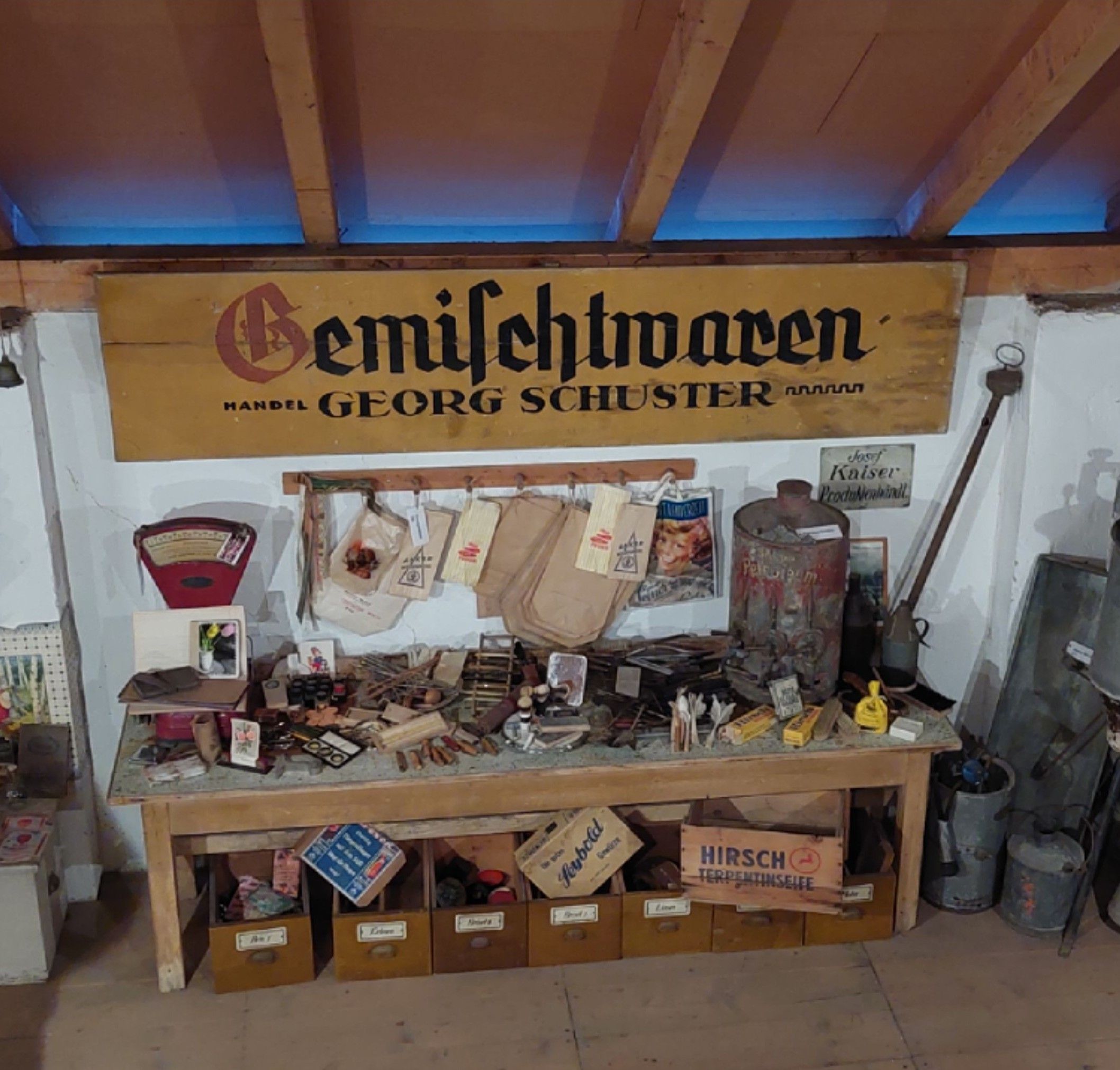 Ein Tisch mit alten Gemischtwaren und einem Schild mit der Aufschrift 'Gemischtwaren Handel Georg Schuster' im Bauernmuseum Kalladorf.