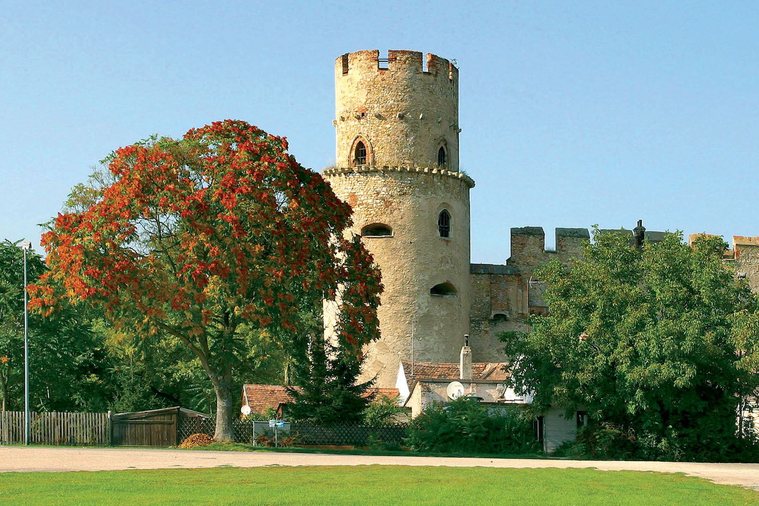 Ein runder Burgturm aus Stein neben einem Baum mit roten Blättern, umgeben von grüner Vegetation und blauem Himmel.