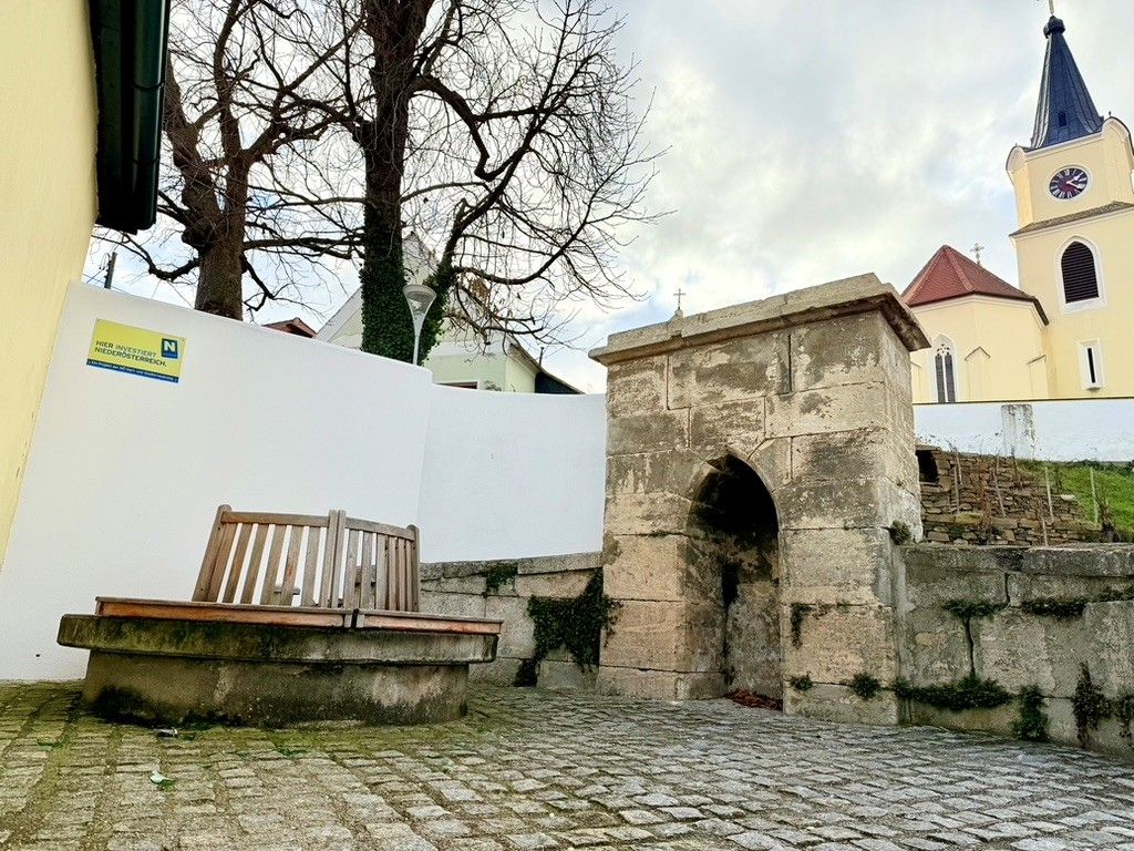 A historic fountain with a wooden bench next to it, in the background a church with a tower and clock.