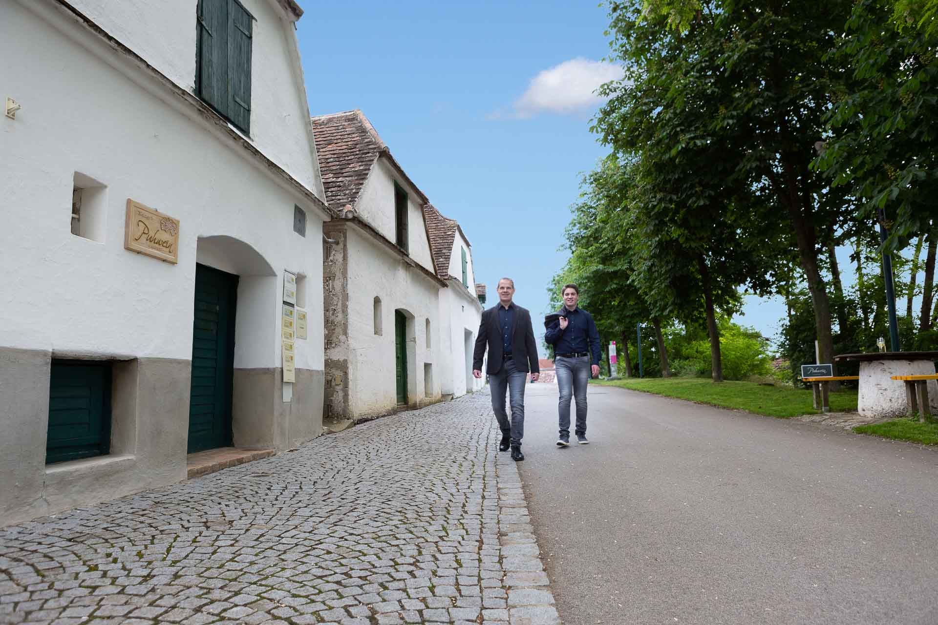 Two men walk along a cobbled street, past white buildings and trees.