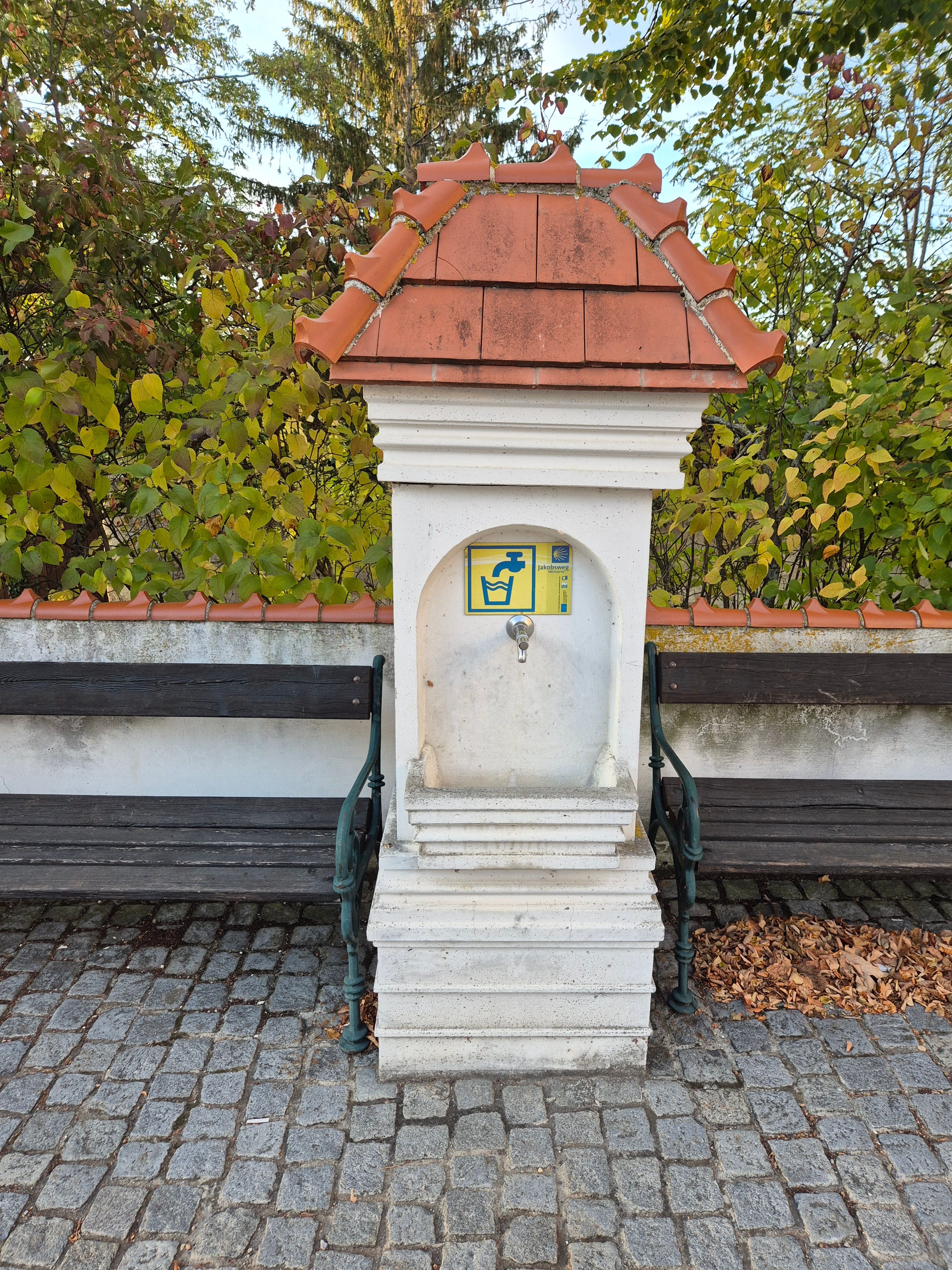 A small fountain with a tiled roof and tap, flanked by two wooden benches on a paved square.