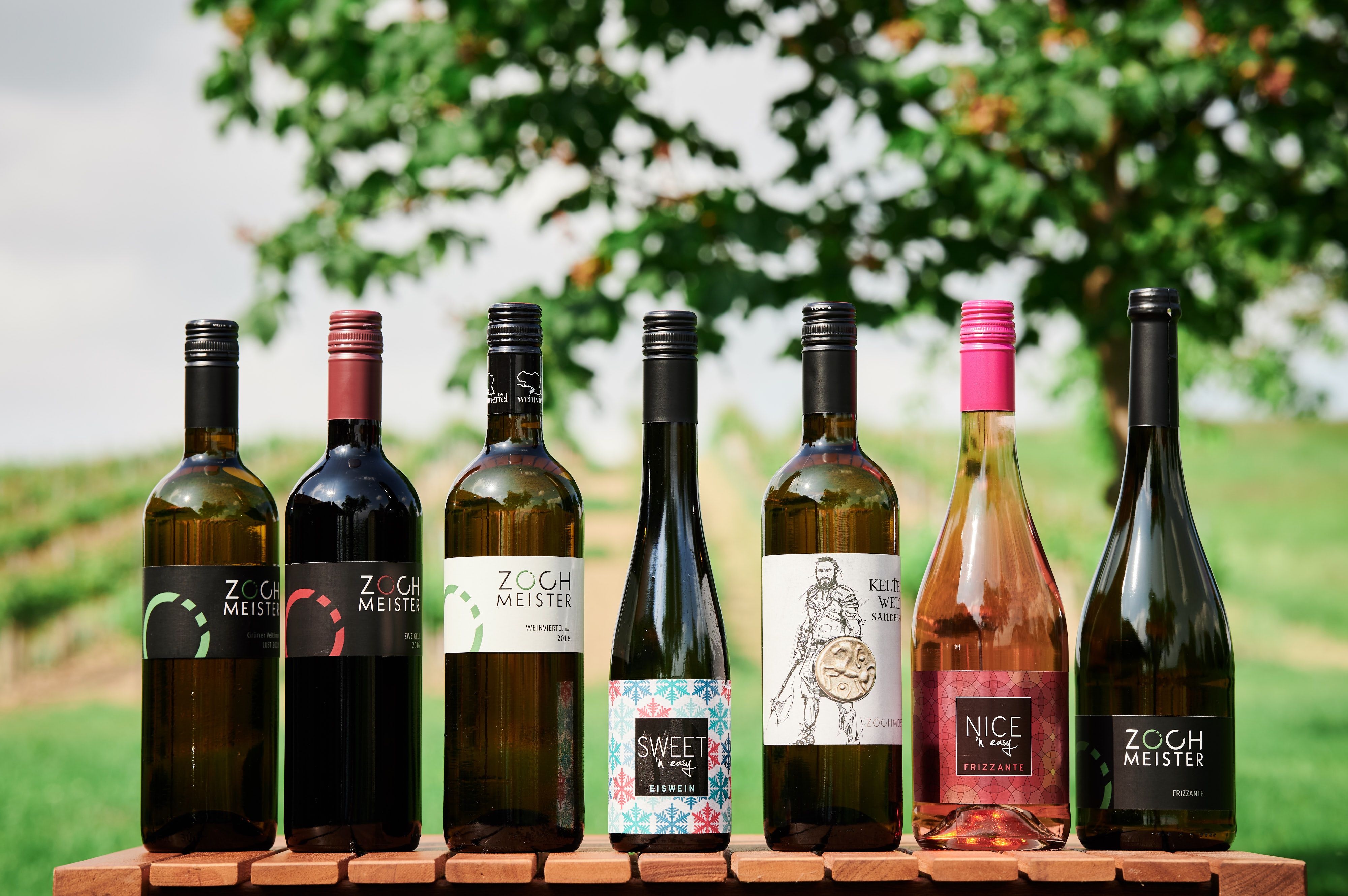 Seven bottles of wine on a wooden table in front of a blurred background with trees and meadow.