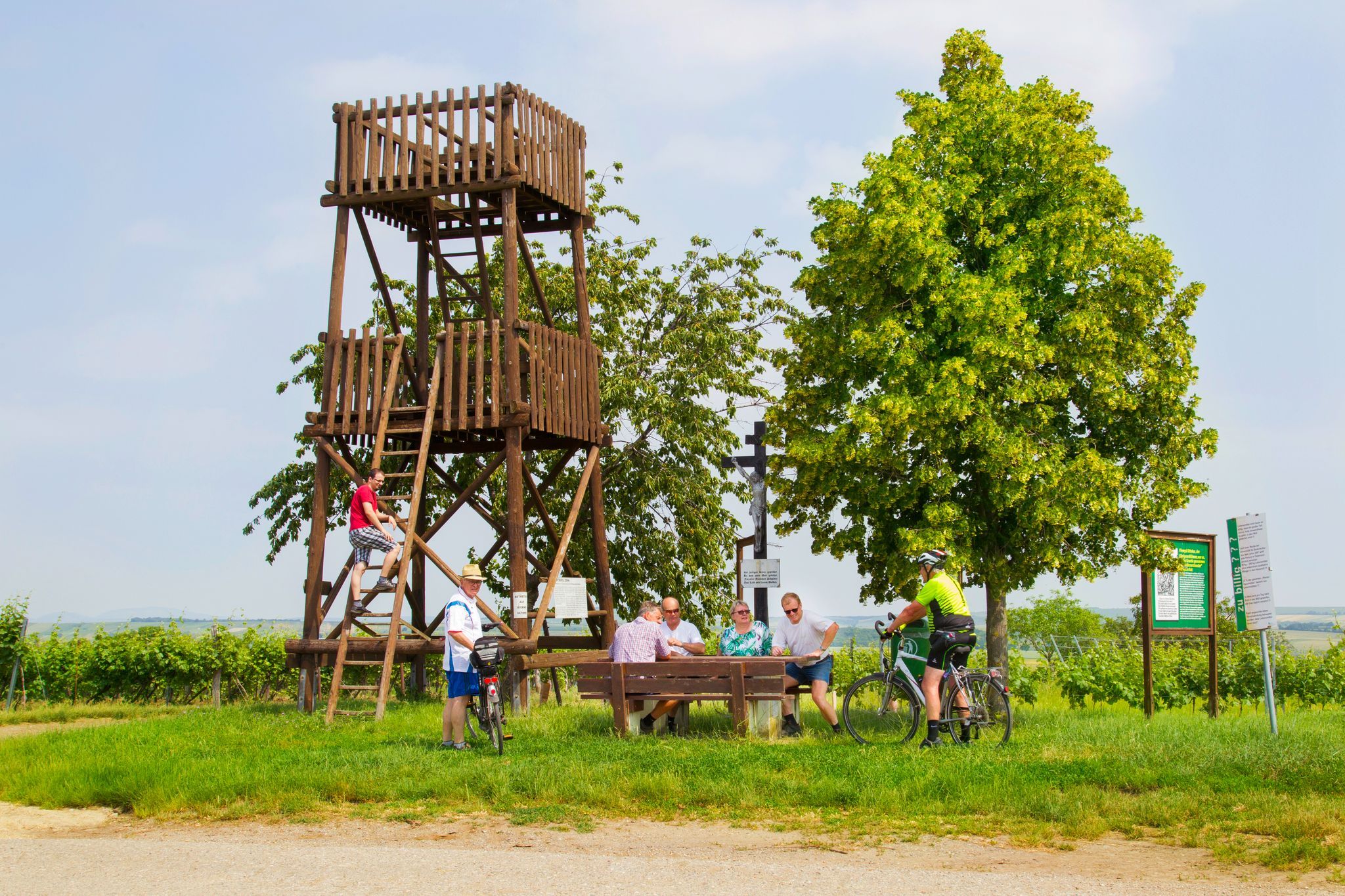 Menschen entspannen an einem Holzturm und Tisch im Freien, umgeben von Bäumen und Schildern.