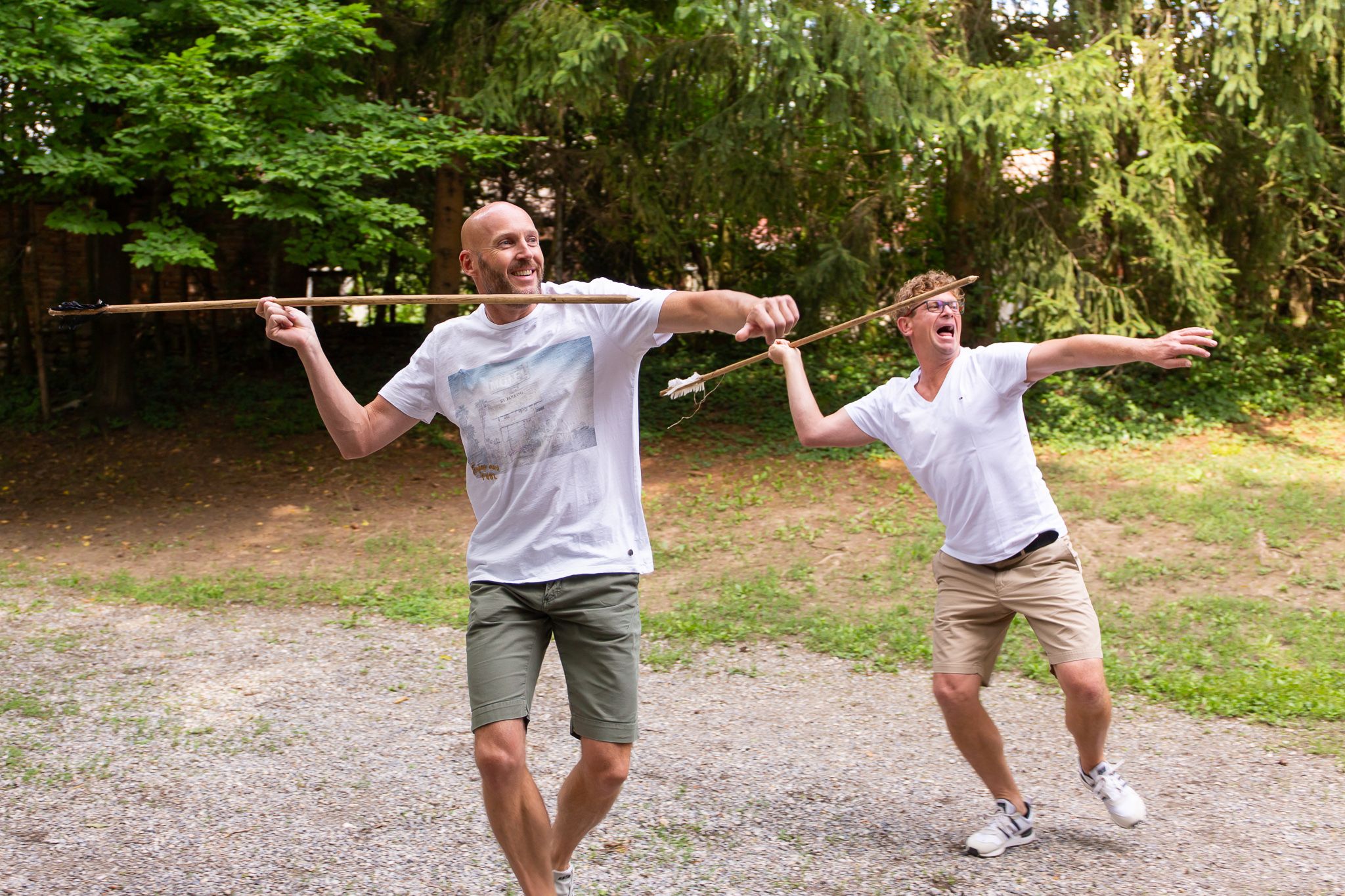 Two men throw spears outdoors, surrounded by trees.