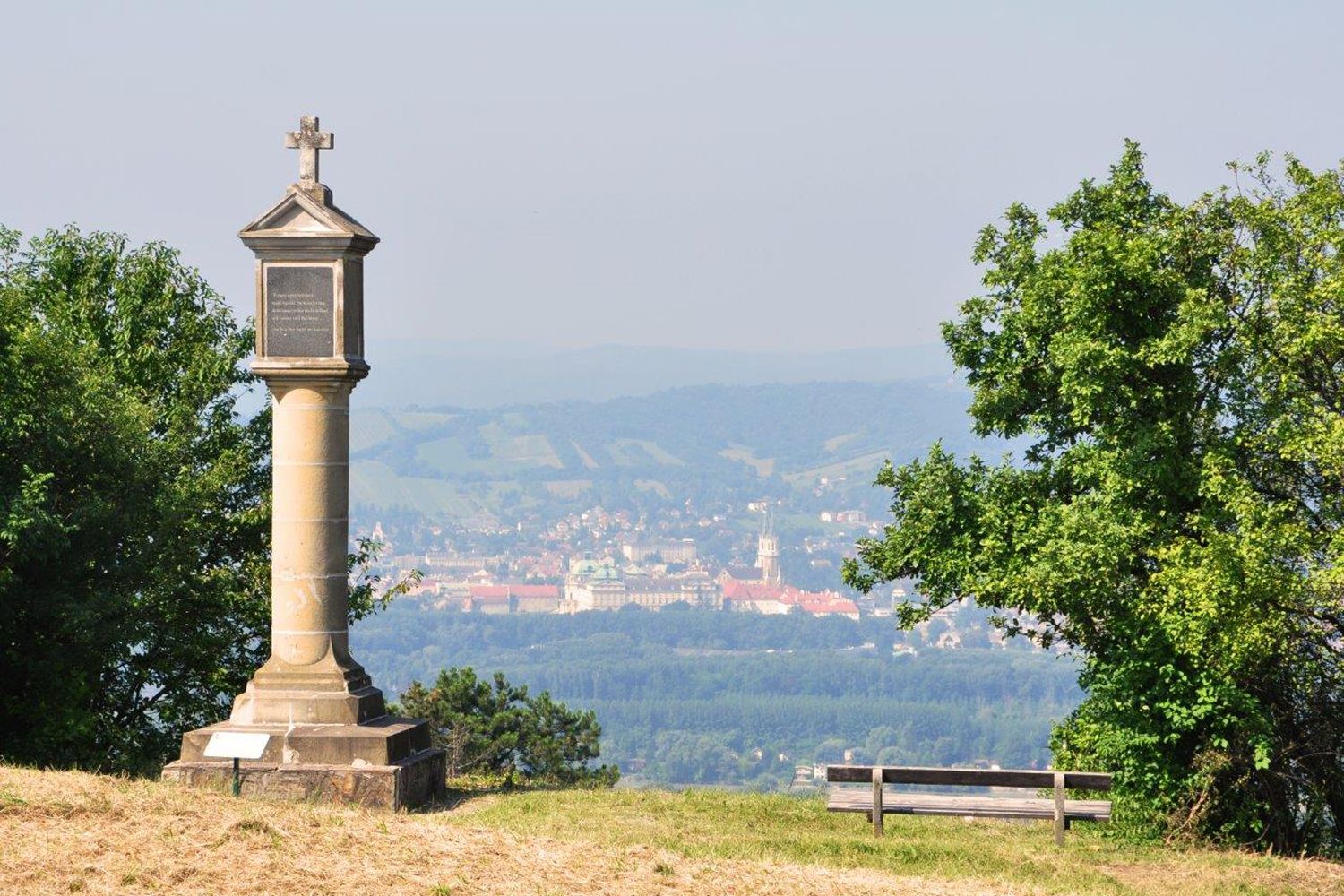 Säule mit Kreuz auf einem Hügel, im Hintergrund eine Stadt und Hügel.