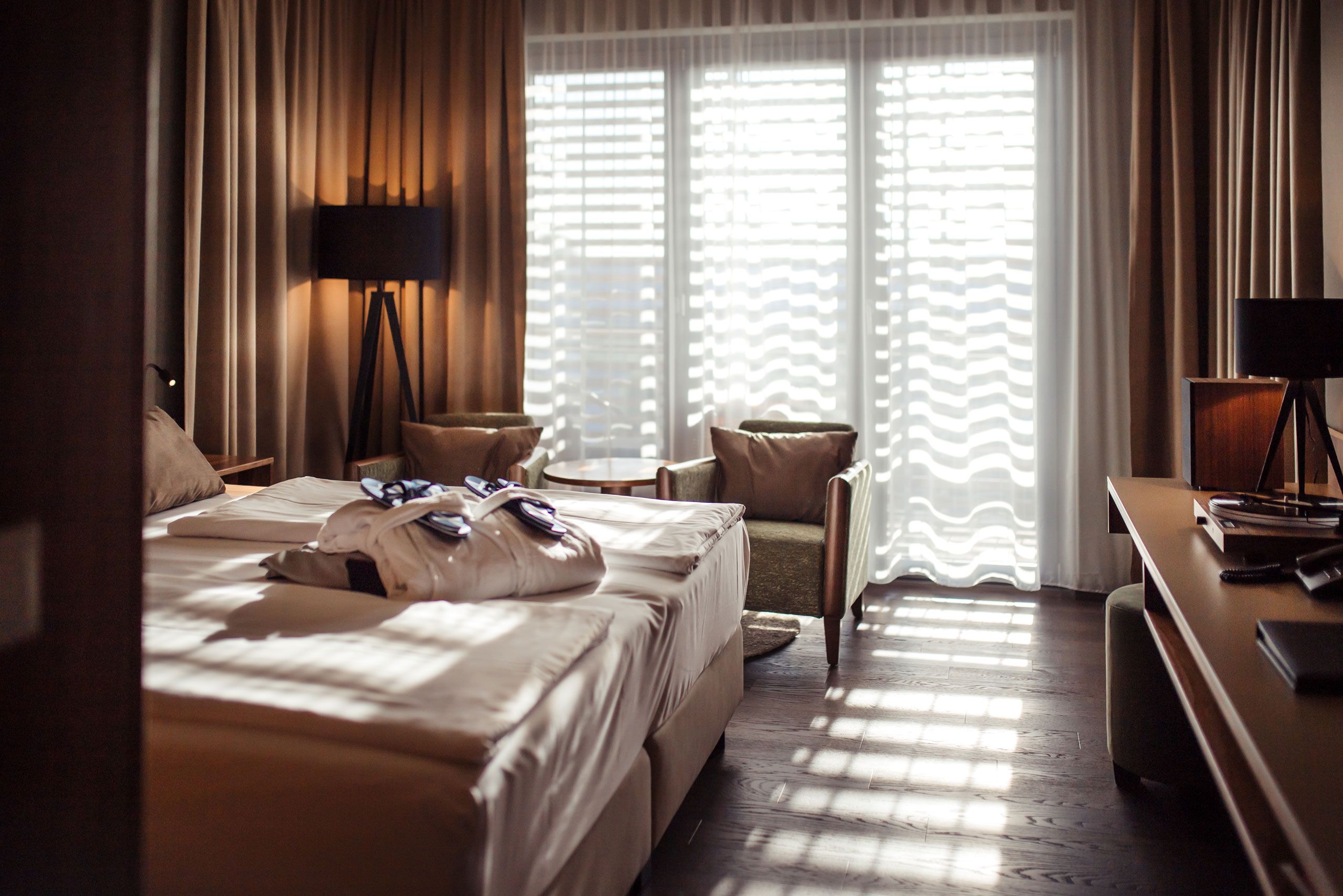 Hotelzimmer mit Doppelbett, Bademänteln, zwei Sesseln und einem Tisch vor einem großen Fenster mit Vorhängen.